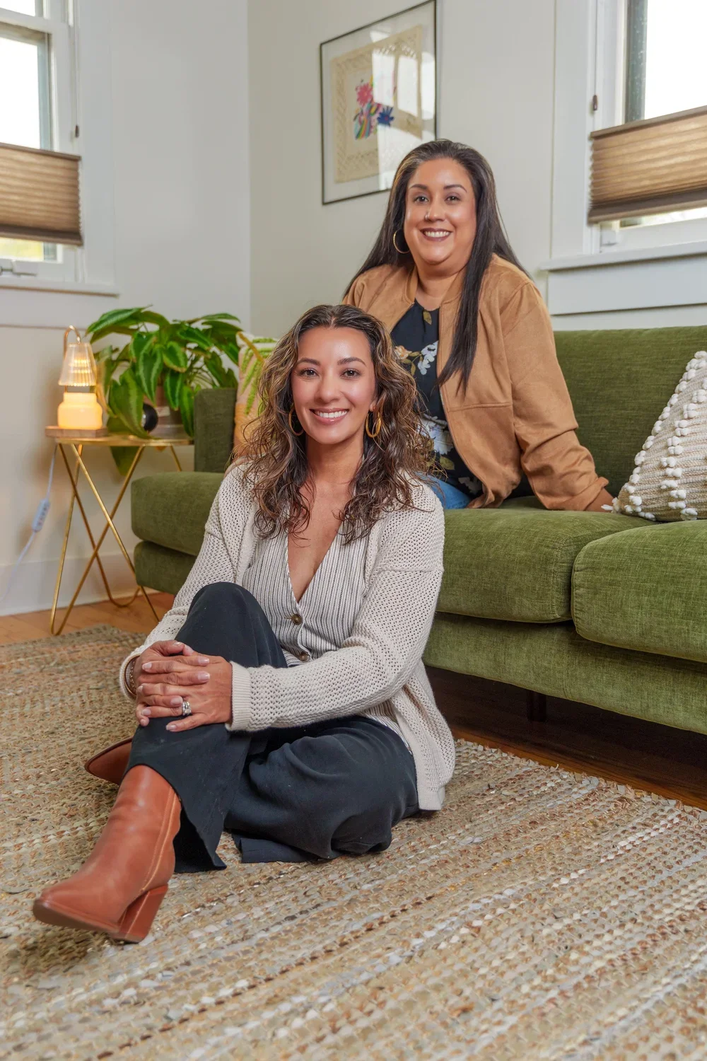 Two women smiling in a cozy living room with a green sofa and a small side table with a lamp and a green plant.