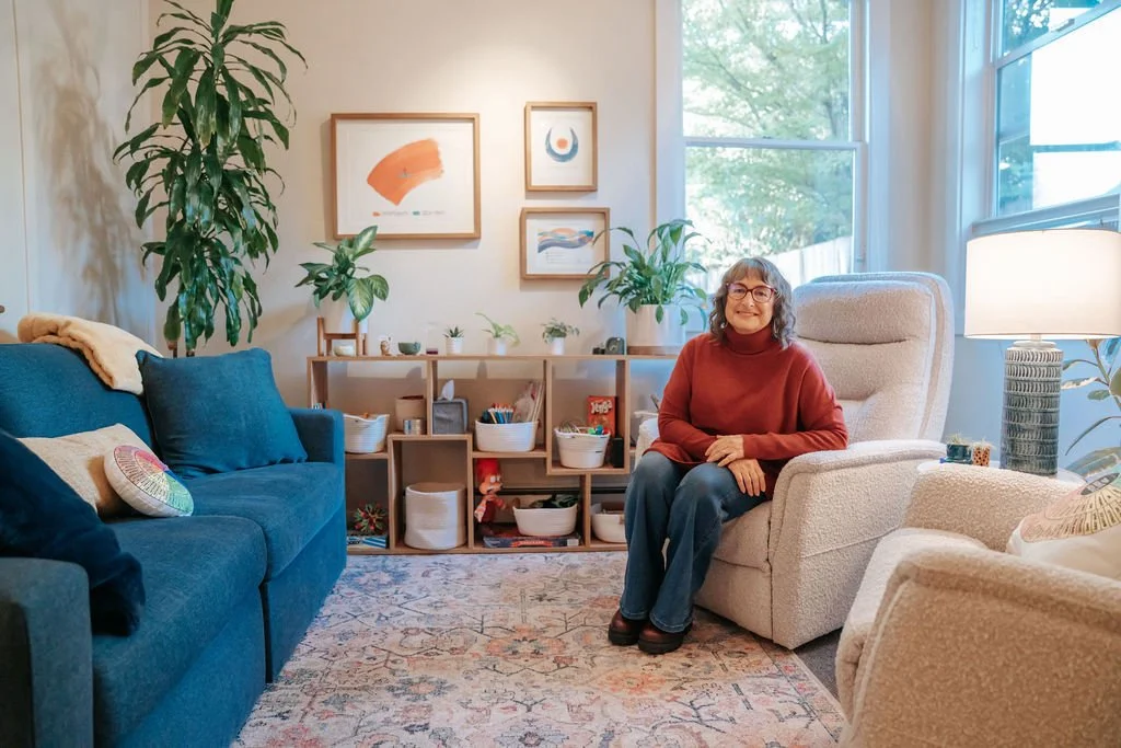 A woman sitting in a beige armchair in a well-lit living room with large windows, a blue sofa, and decorative plants. The room has artwork on the wall and a bookshelf with various items.