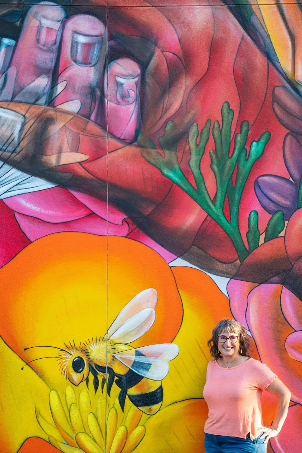 A woman with glasses and curly hair smiling in front of a colorful mural featuring a bee, large flowers, and greenery.
