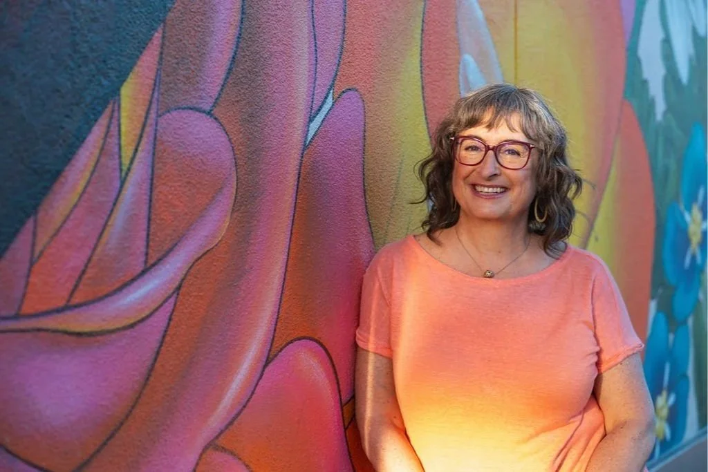 A smiling woman with curly gray hair and glasses standing next to a colorful mural of a hand.