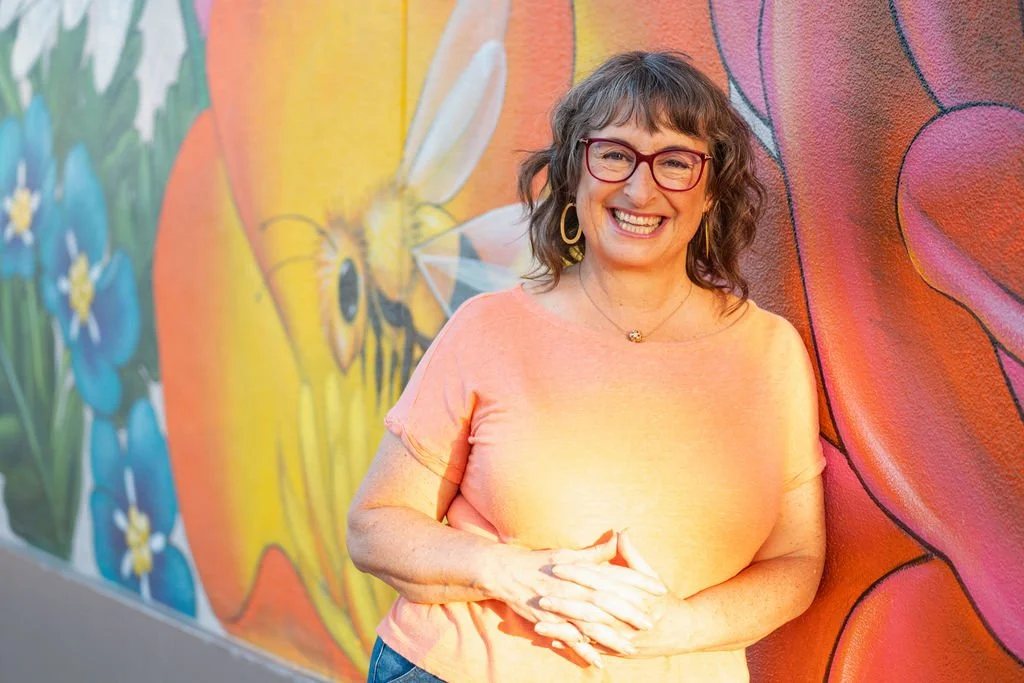 A woman with shoulder-length curly gray hair, red glasses, and a big smile, standing in front of a colorful mural with a bee, blue flowers, and pink petals.
