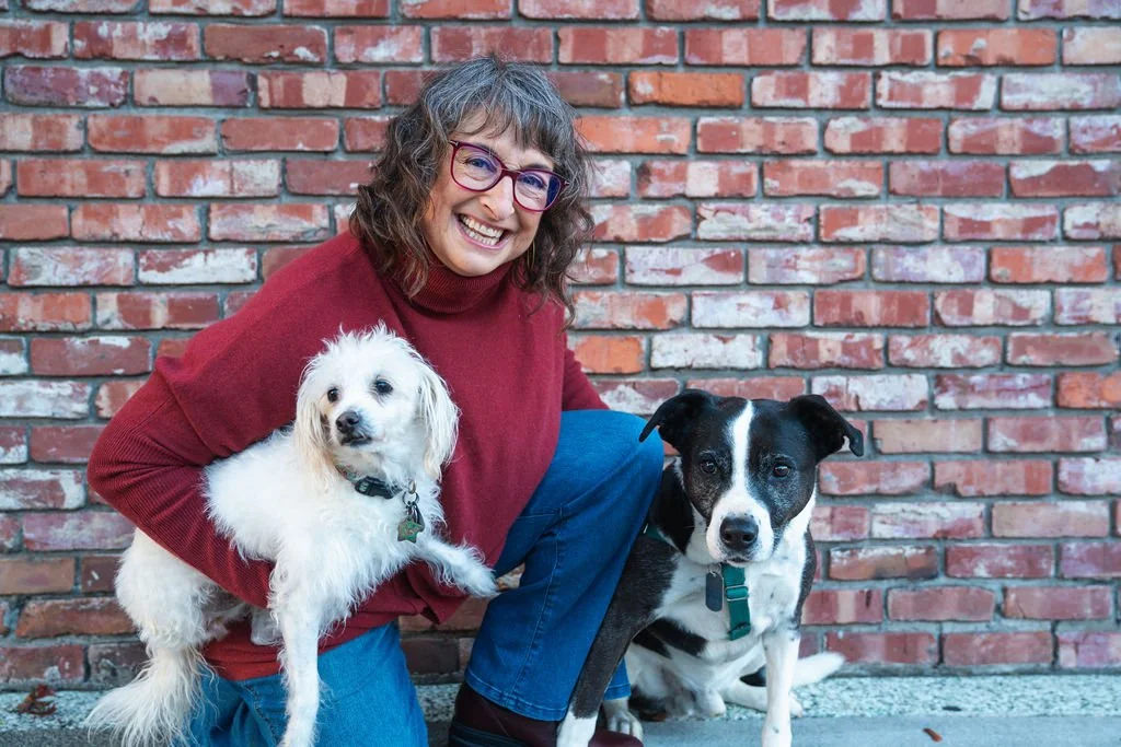 A woman with glasses, wearing a red sweater, smiling while squatting outdoors in front of a brick wall, holding a small white dog with floppy ears and a black collar. Next to her, a black and white dog with a teal collar is sitting on the ground.