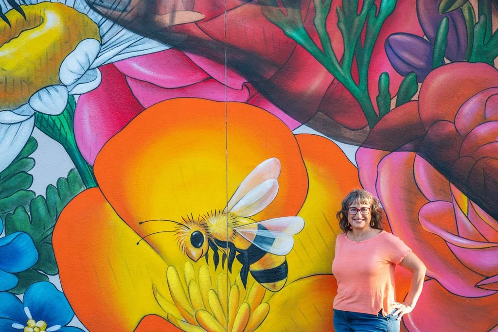 A woman wearing glasses and a pink shirt stands in front of a large, colorful mural of a bee and flowers.