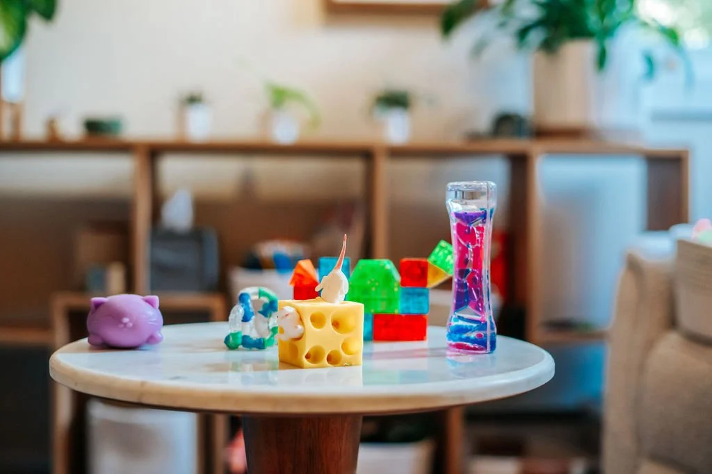 A small table with colorful toy building blocks, a purple pig figurine, and a tall, slender water toy with pink and purple liquid inside, in a cozy living room with shelves and plants in the background.