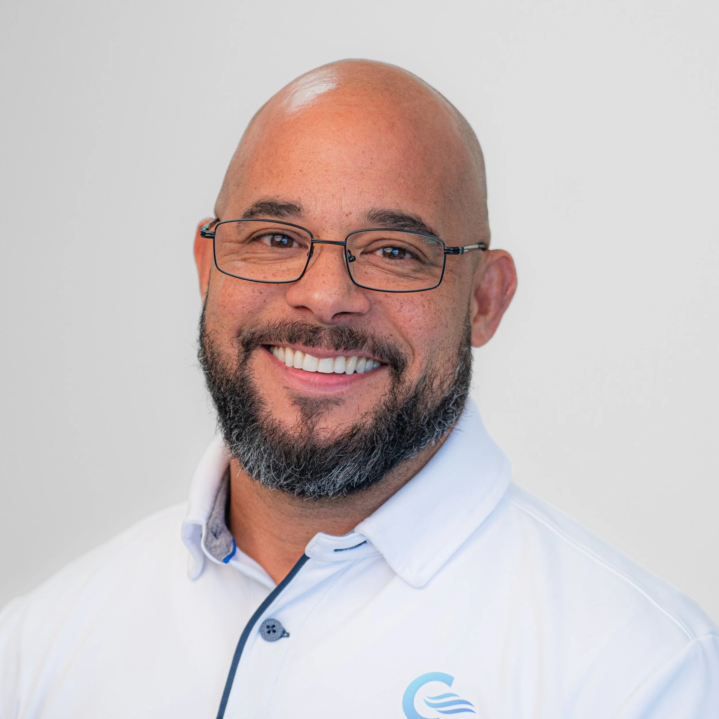 A smiling man with glasses, a beard, and a bald head, wearing a white collared shirt with a logo, against a plain white background.