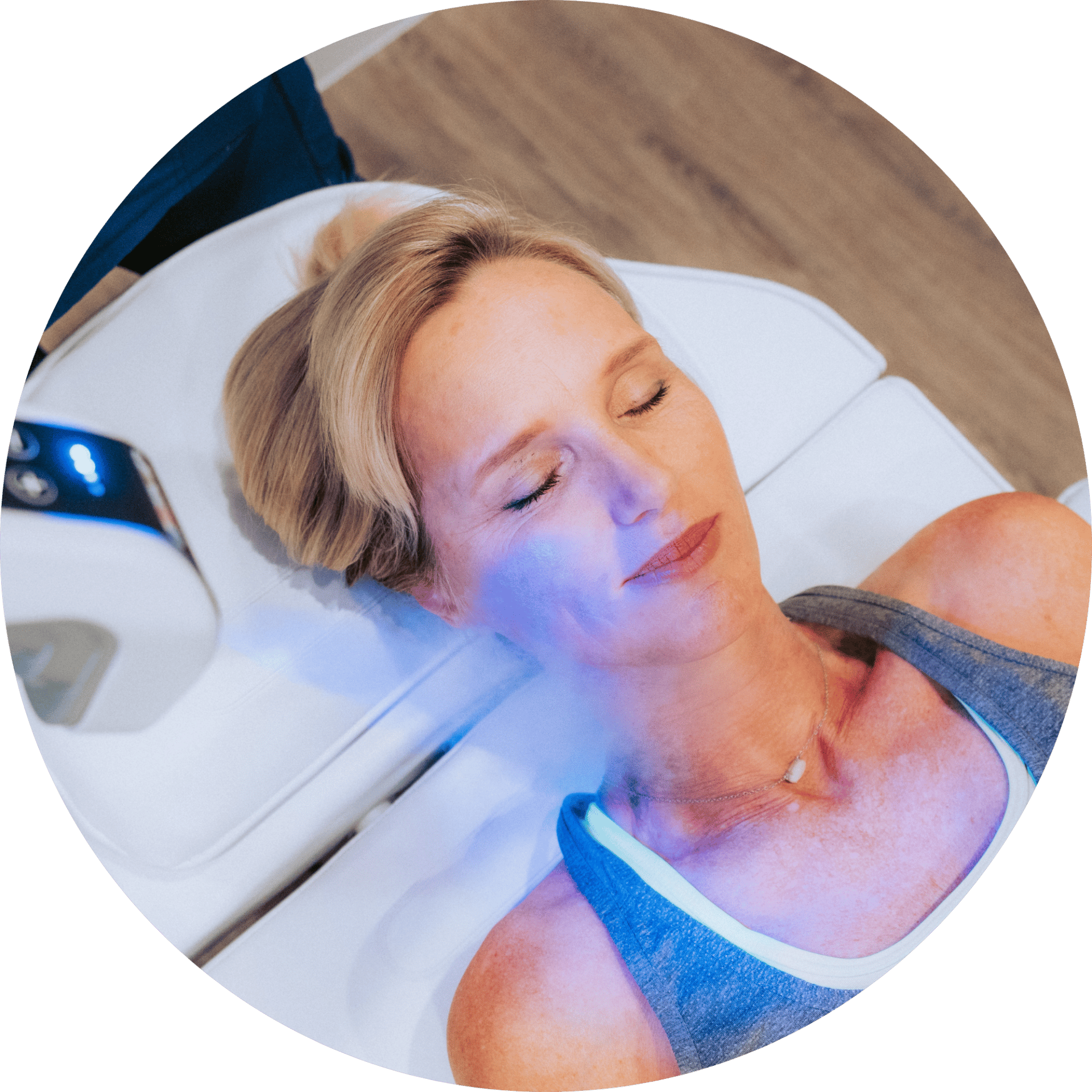 A woman with blonde hair receiving red light therapy on her face while lying on a white treatment bed.