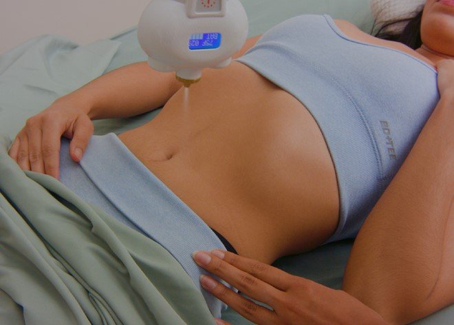 A woman lying down on a medical bed during an ultrasound exam. A healthcare professional is performing the ultrasound on her abdominal area with a device that has a blue digital display.