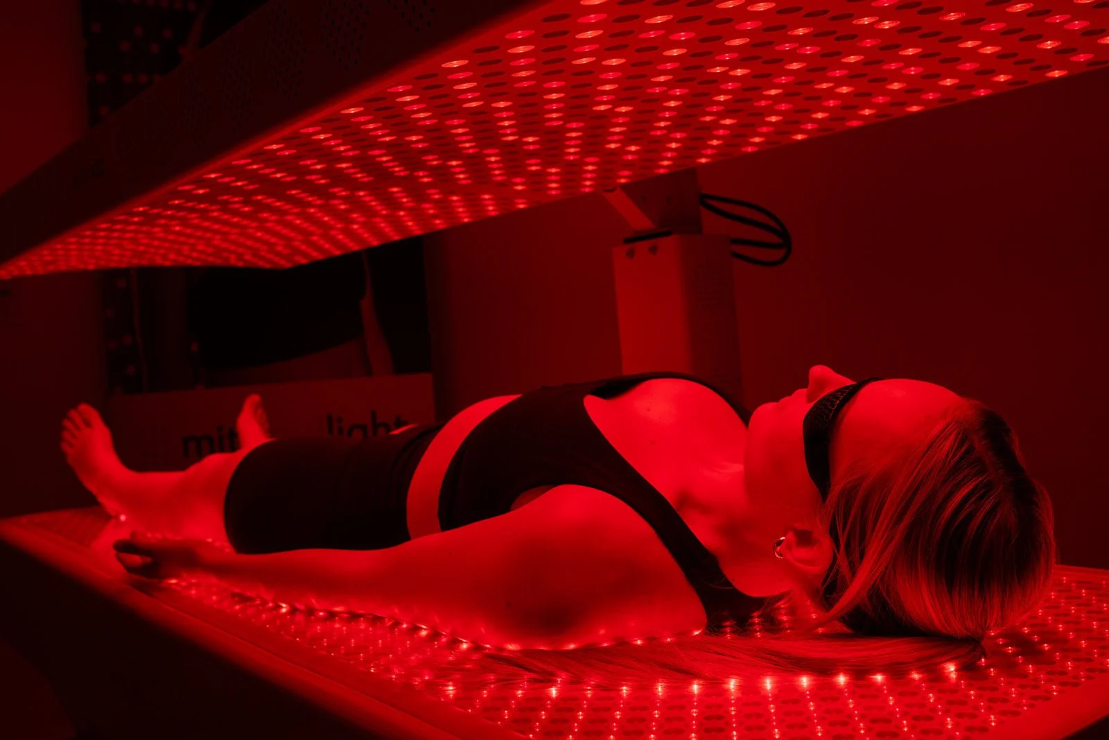 A woman lying on a purple light therapy bed with red LED lights, wearing black athletic wear and eye protection in a dark room.