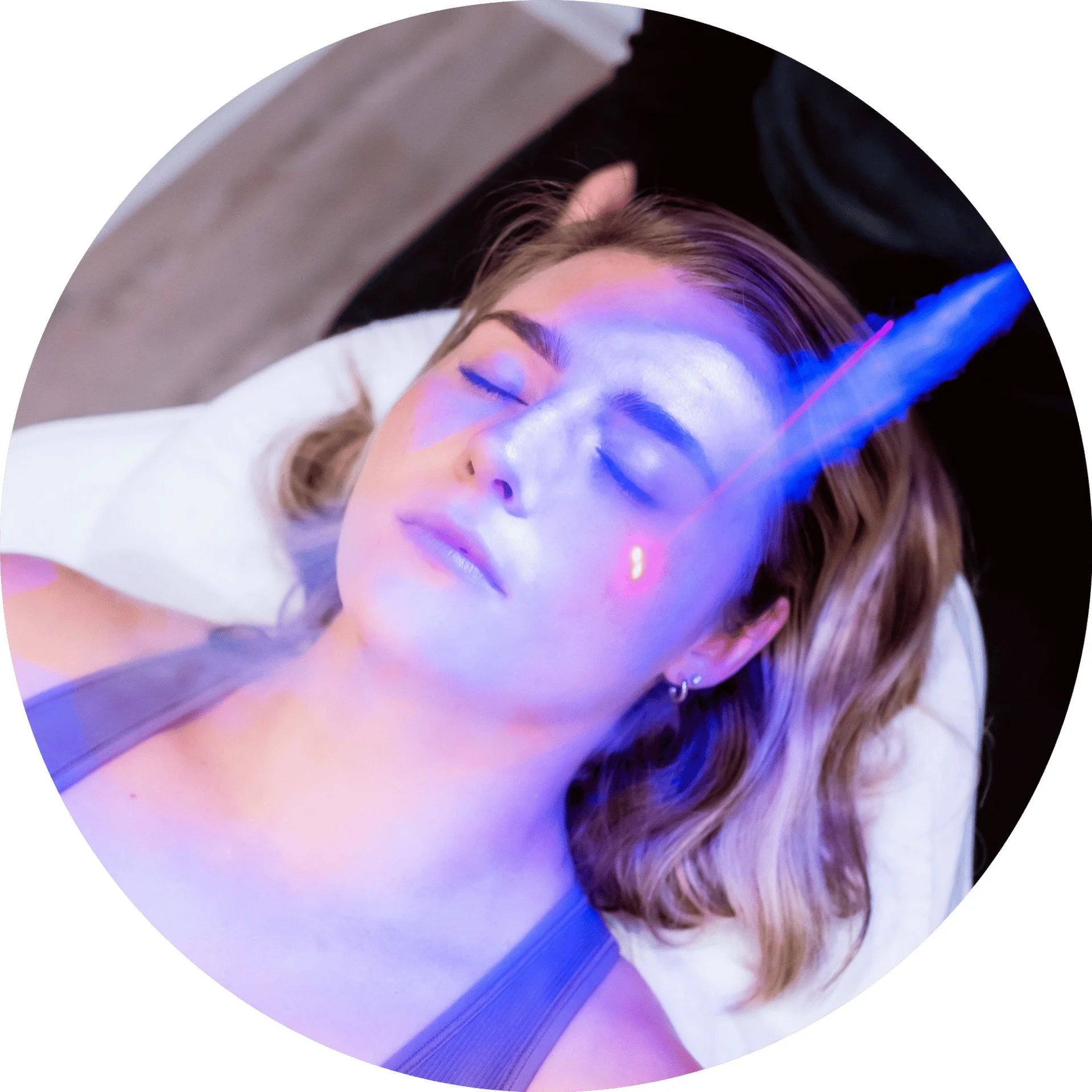 A woman with light brown hair and earrings lying down with her eyes closed receives a laser treatment on her face, with a beam of blue and pink light projected onto her skin.