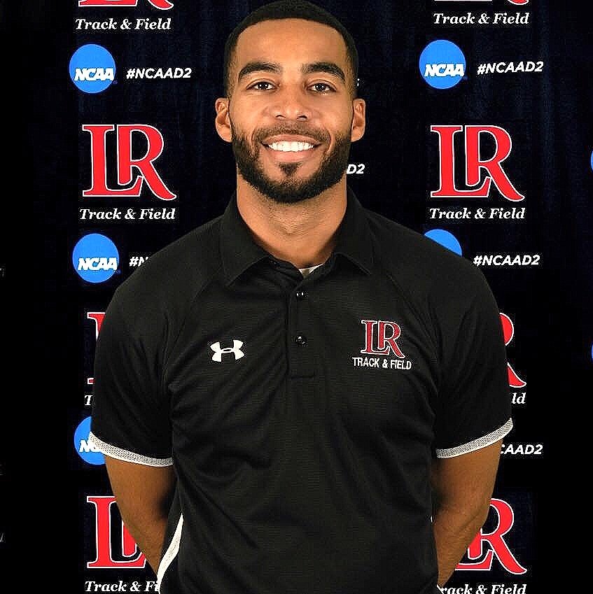 A man with short hair and beard, smiling, wearing a black collared shirt with the LR Track & Field logo and an Under Armour logo, standing in front of a backdrop with LR Track & Field and NCAA logos.