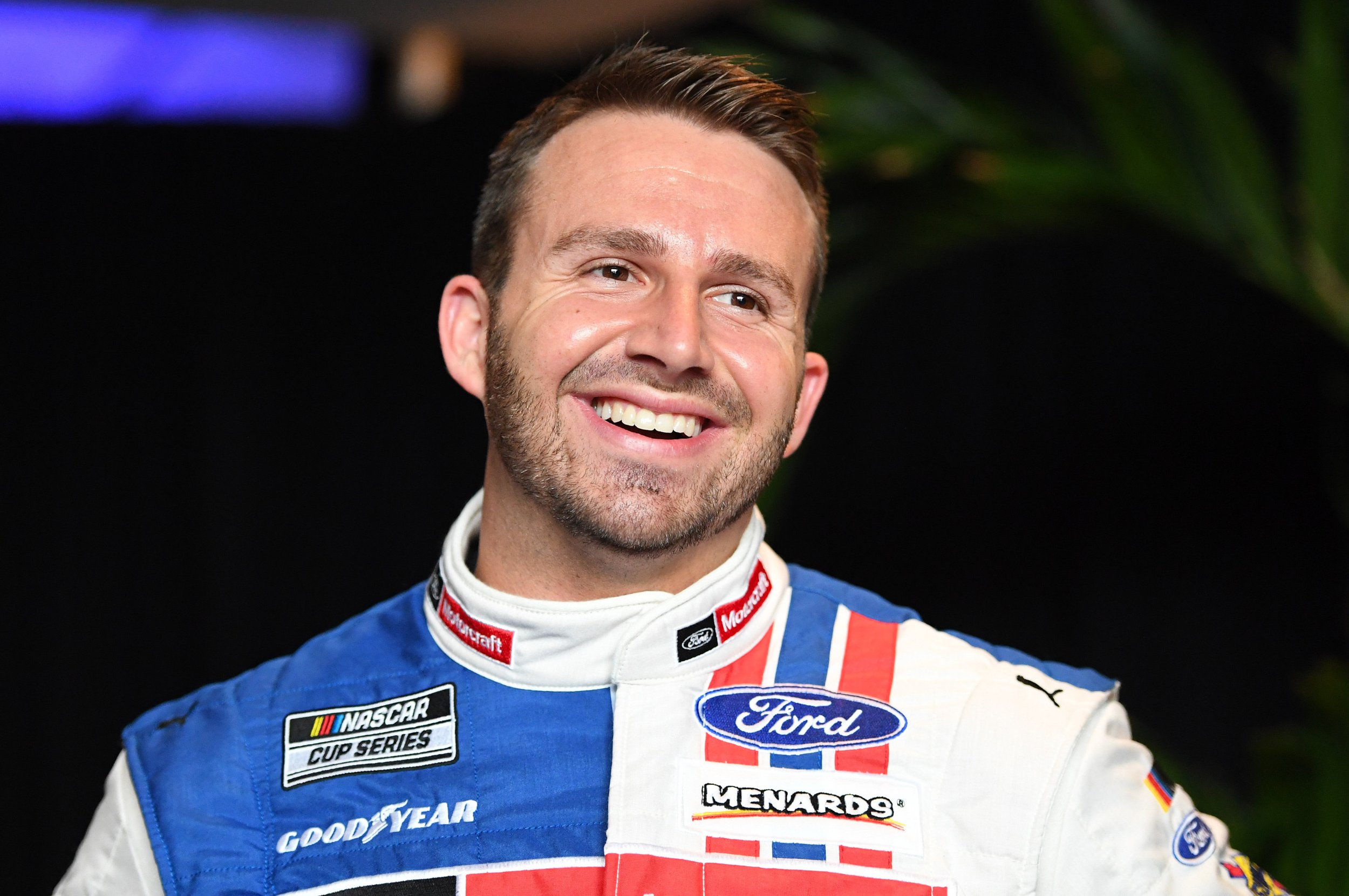 Smiling male race car driver in racing suit with NASCAR and Ford logos.