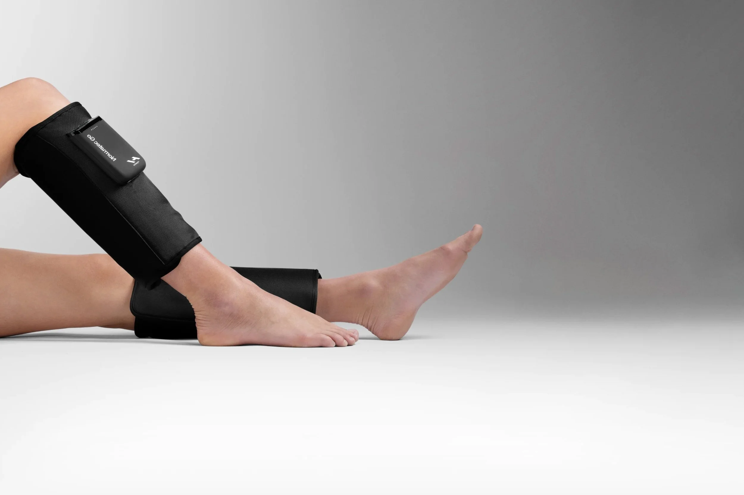 Legs in a stretch position with medical devices attached to the lower leg and thigh, on a gray background.