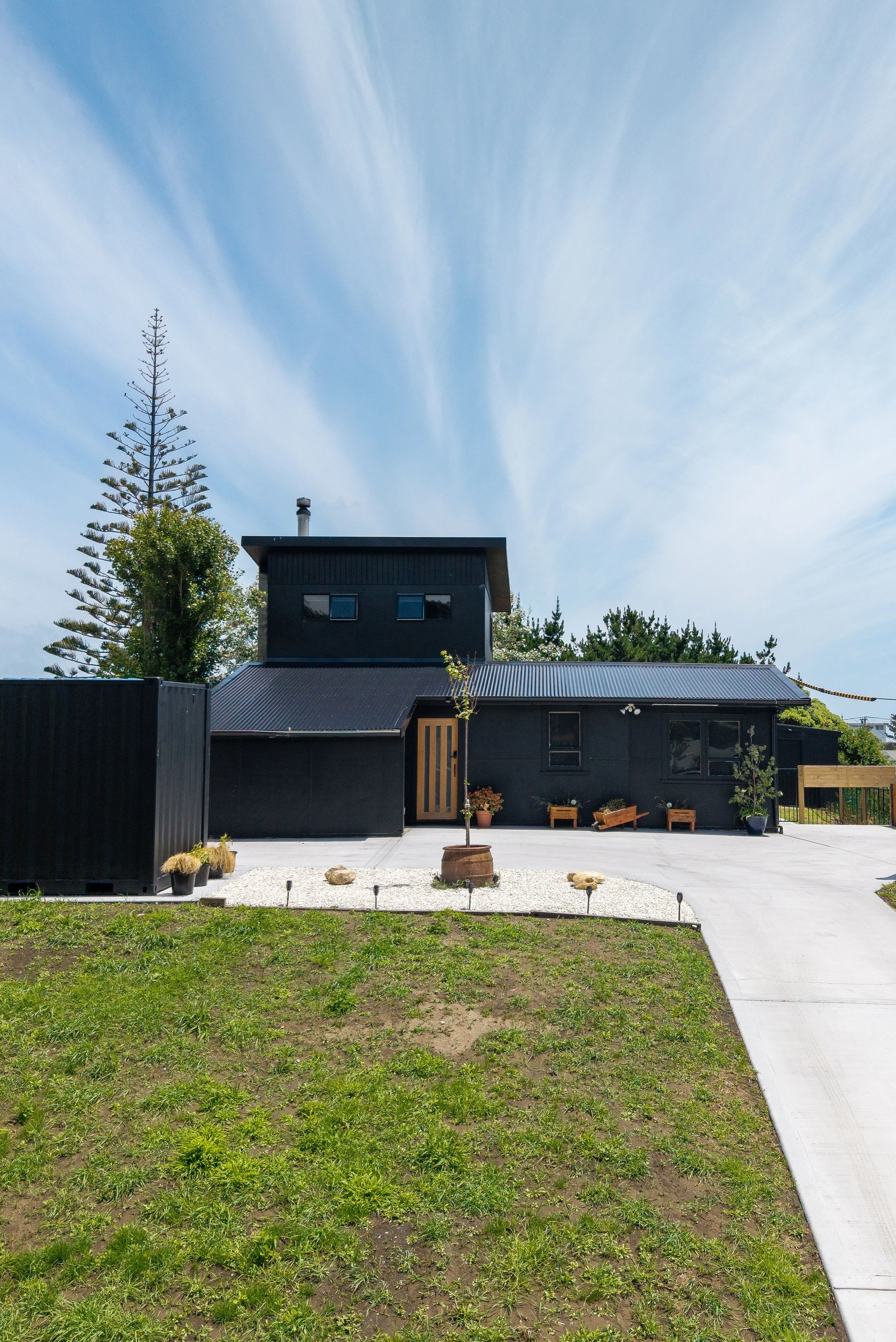 A modern black house with potted plants and benches in a yard under a partly cloudy blue sky.