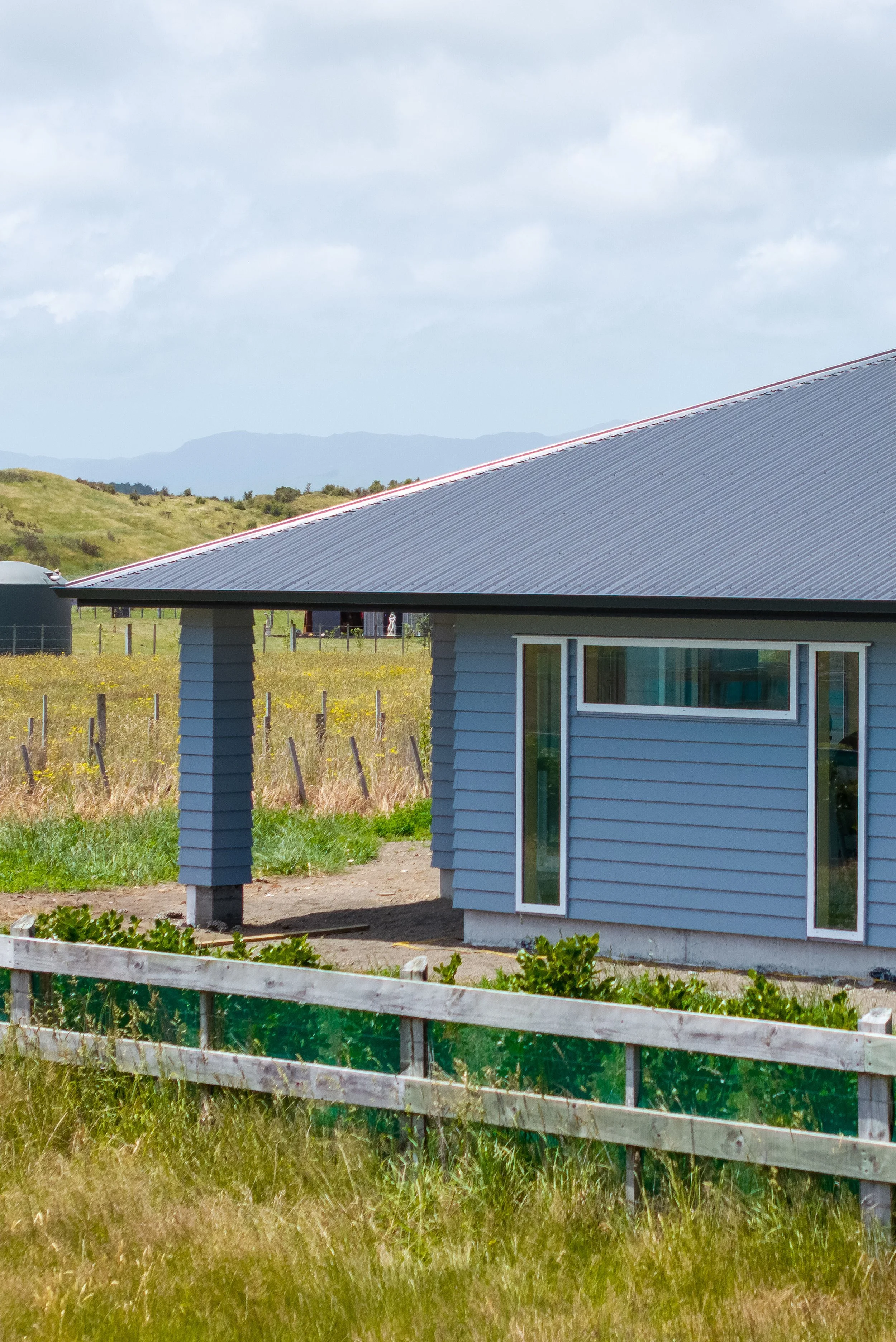 A country house with blue siding and a metal roof in a rural area, surrounded by green fields and fencing.