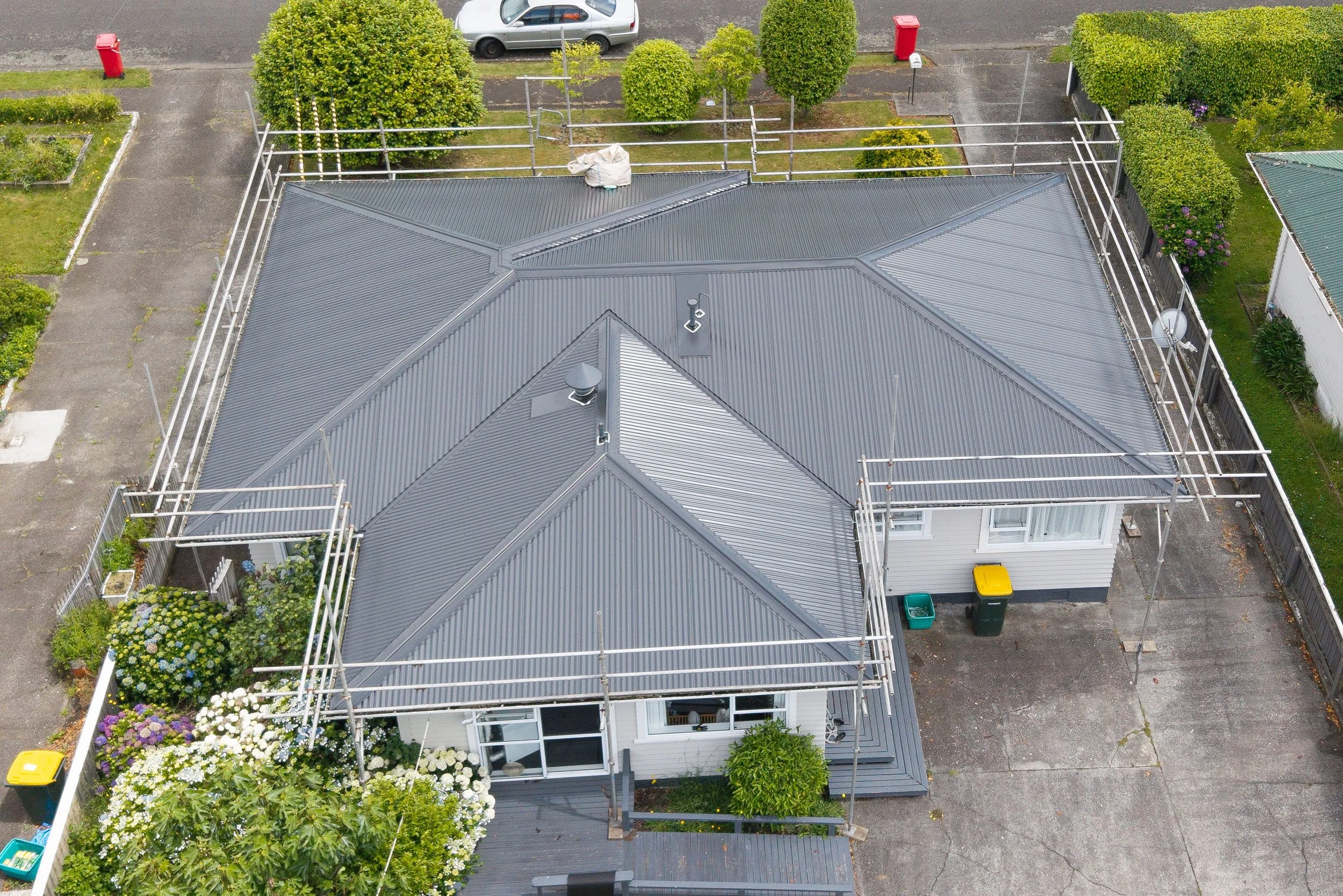An aerial view of a house with an intricate metal roof under construction, surrounded by a fenced yard with colorful bushes and trees. There are two cars parked on the street and a few garbage bins on the driveway.