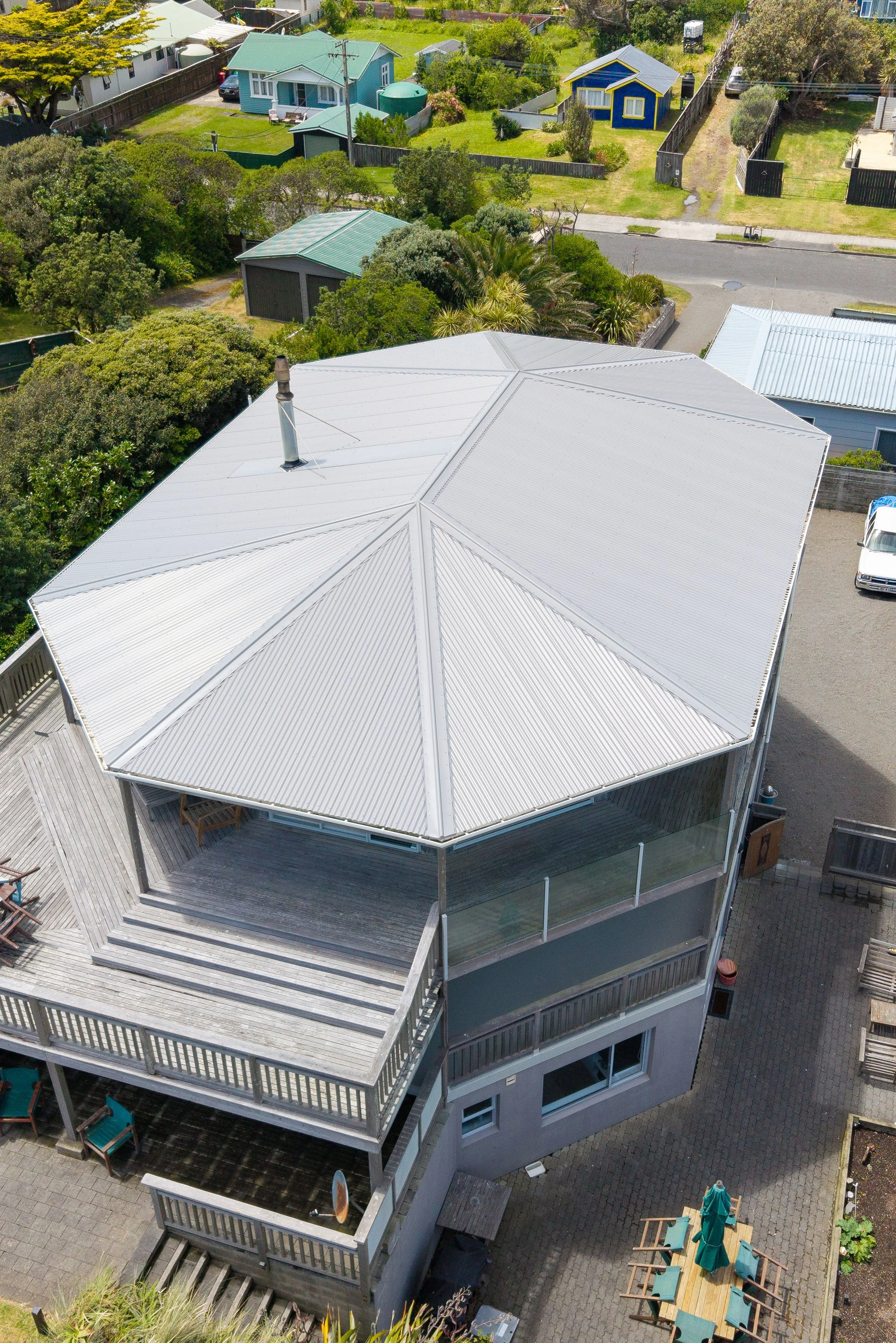 A large house with a multi-section gray metal roof, multiple decks, and outdoor furniture, surrounded by neighboring houses, trees, and a street.