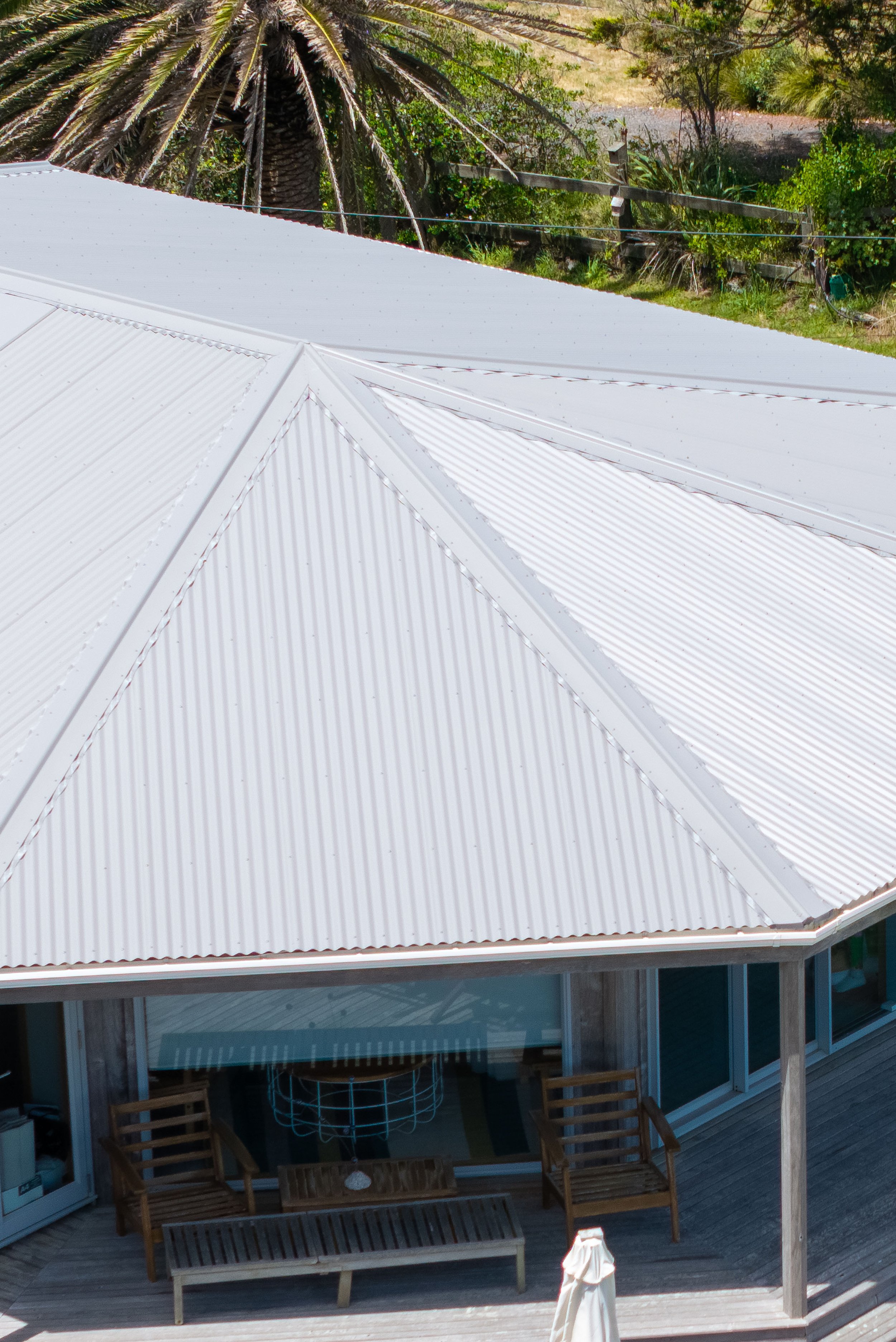 A white metal roof on a house or building with wooden deck and outdoor furniture, with trees and greenery in the background.
