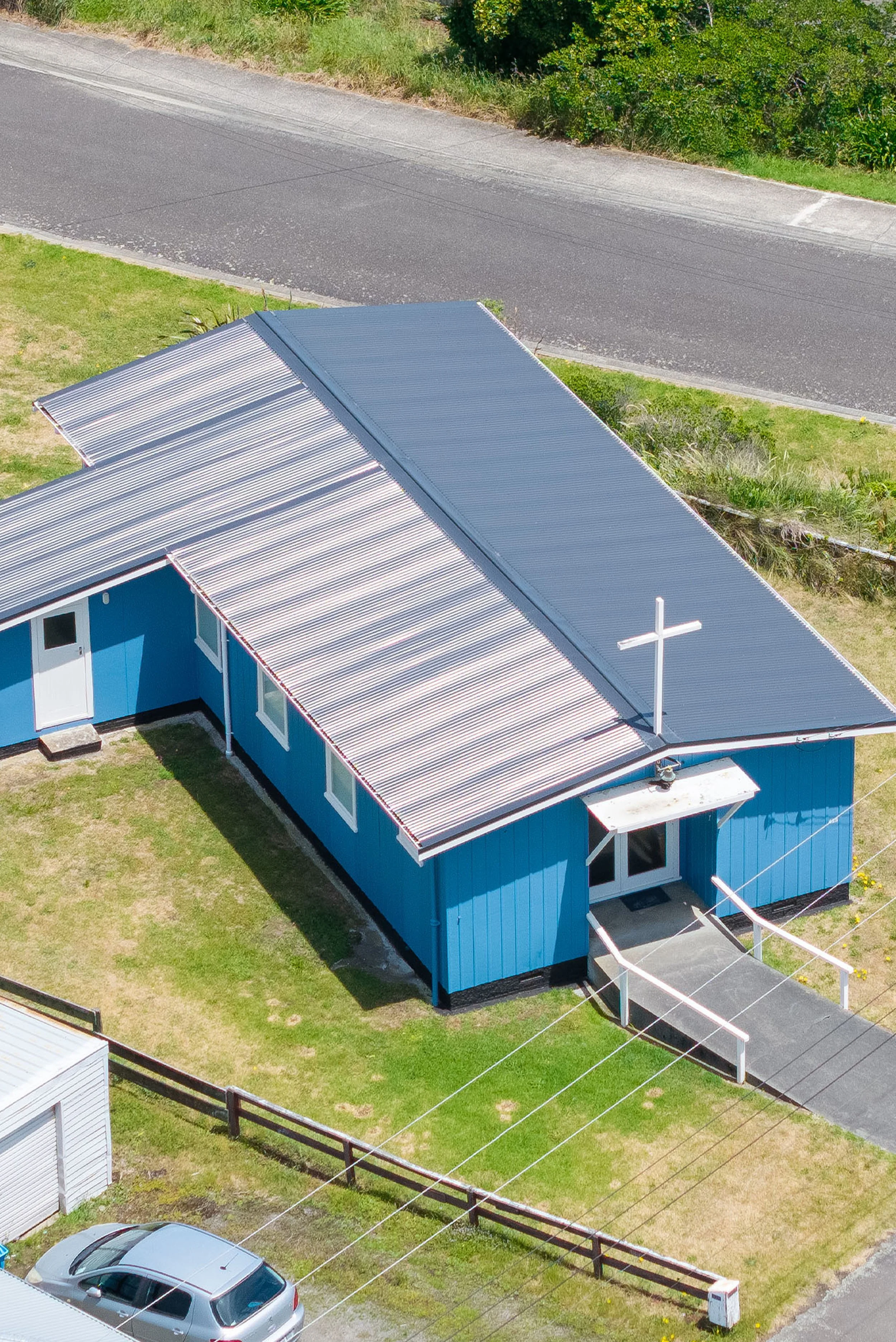 Aerial view of a blue church building with a white cross on the roof, situated on a grassy lot next to a church parking lot.