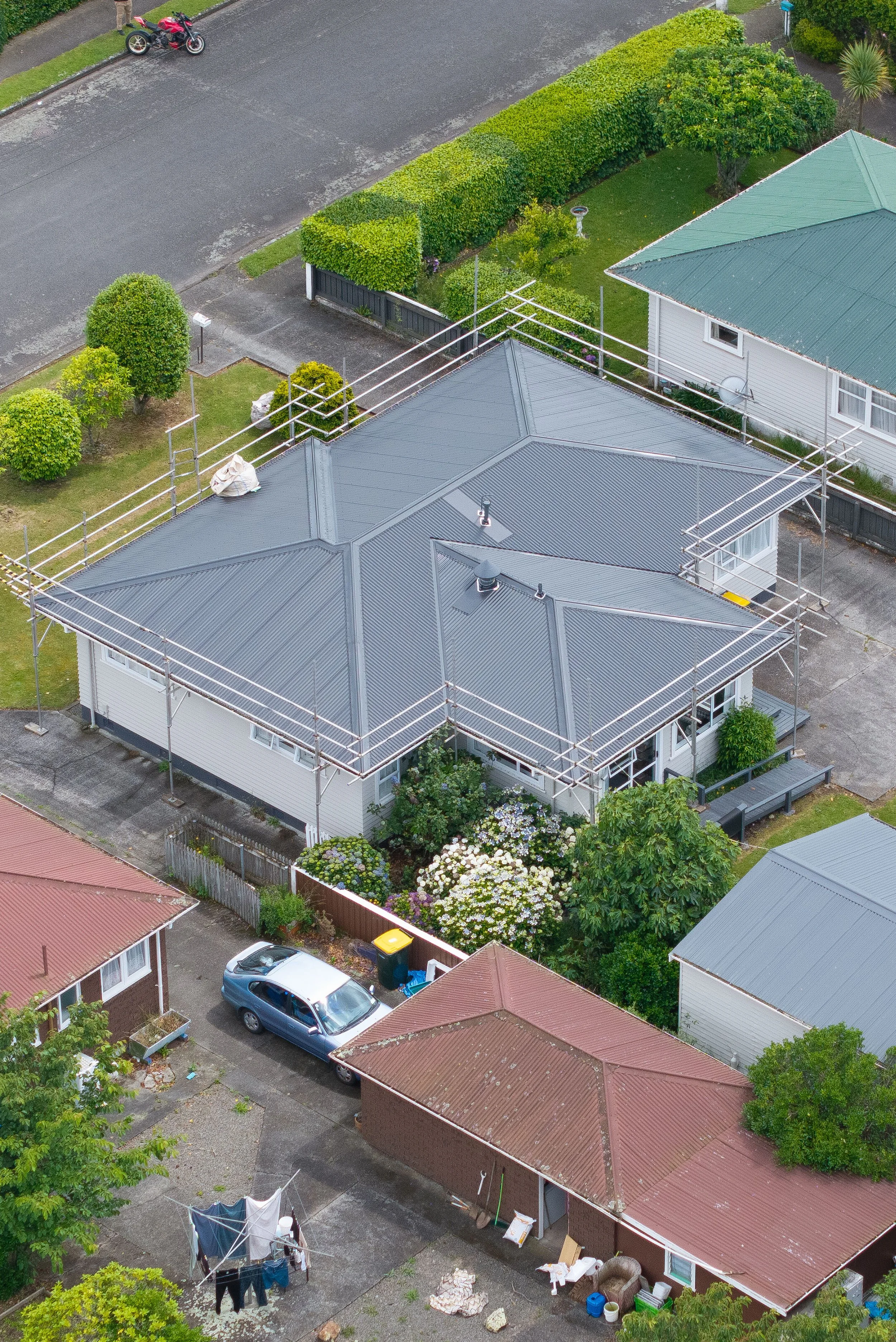 Aerial view of a house with a gray metal roof surrounded by greenery, scaffolding around the roof, neighboring houses, a parked car, bushes, and a clothesline with hanging laundry.