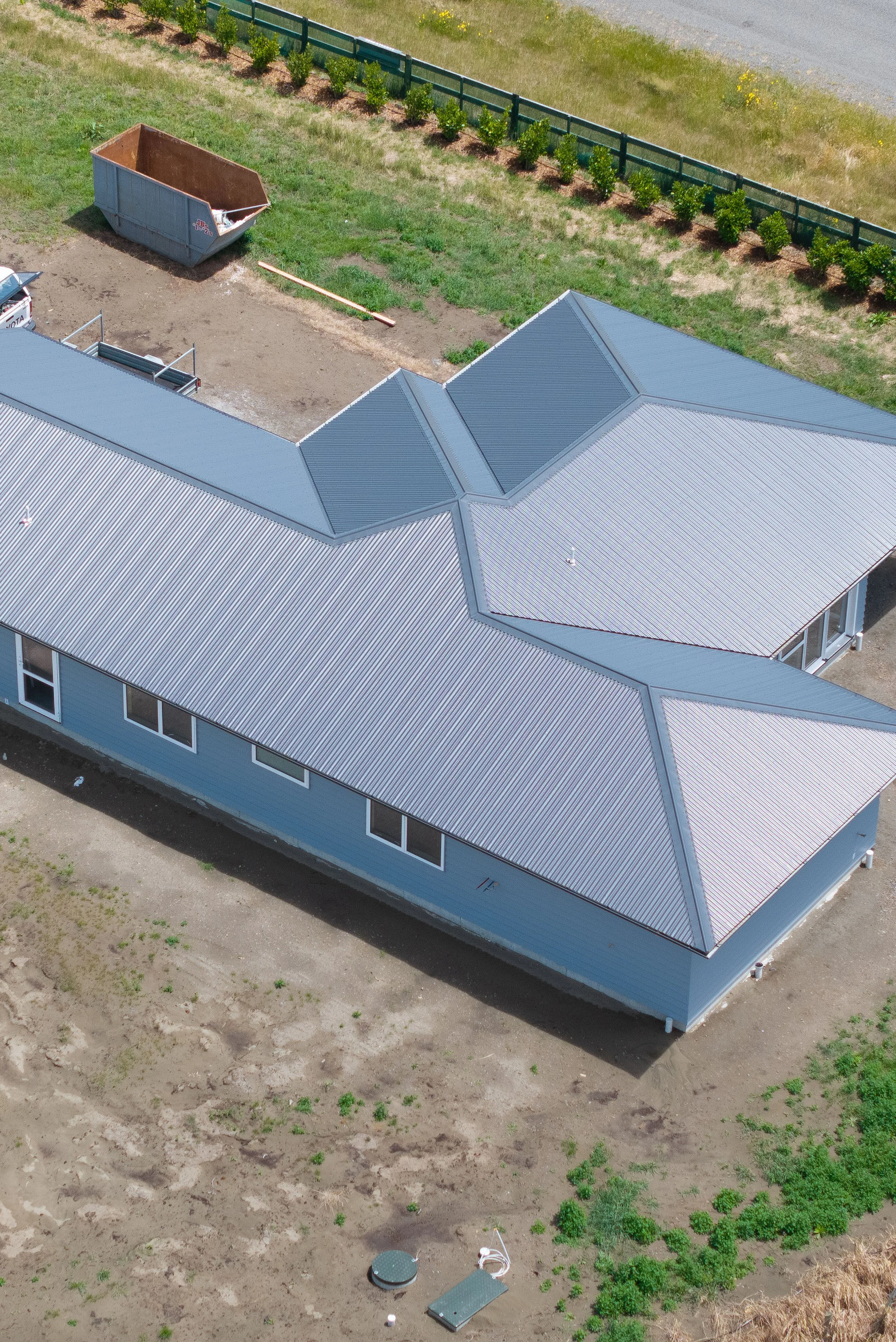 Aerial view of a single-story house with a metal roof, surrounded by a yard with patches of grass and dirt, a row of small trees along a fence, and construction equipment nearby.