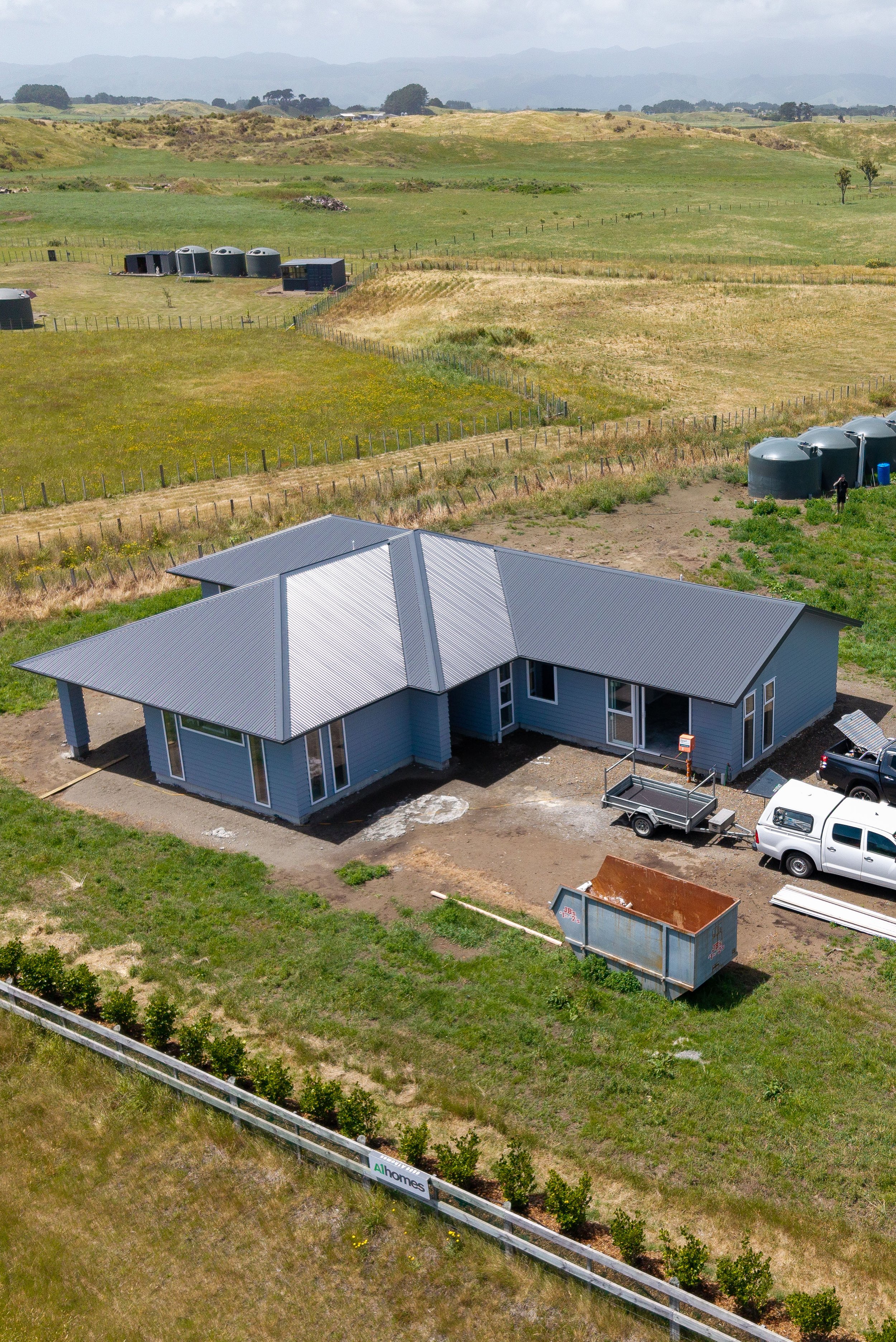 A house under construction with a metal roof, construction vehicles, and a fenced yard, situated in a rural area with open fields and rolling hills in the background.