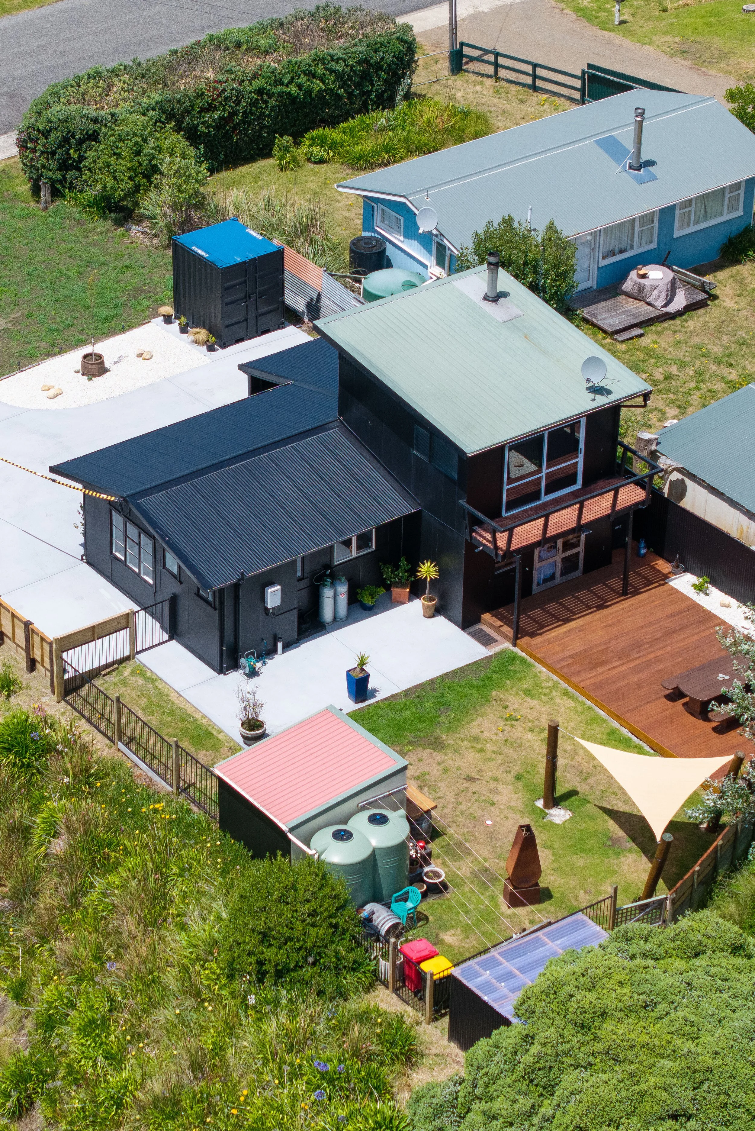 Aerial view of multiple houses with green and metal roofs, fenced backyard with wooden deck, patio furniture, garden, and storage shed.