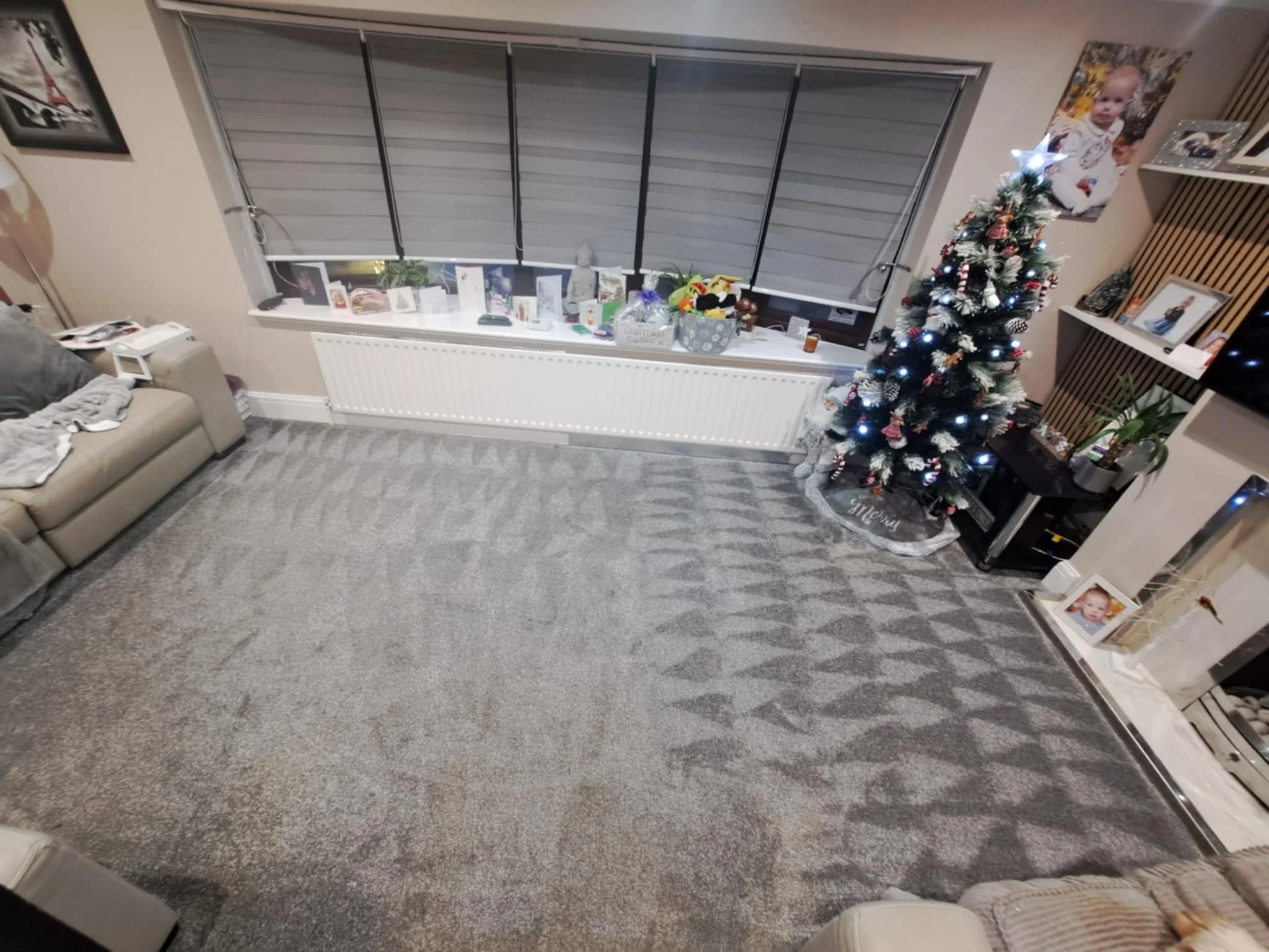 Living room with gray carpet, beige couch, decorated Christmas tree with ornaments and lights, window with closed blinds, various Christmas cards and gifts on the window sill, framed photographs and a lamp on a white shelf.