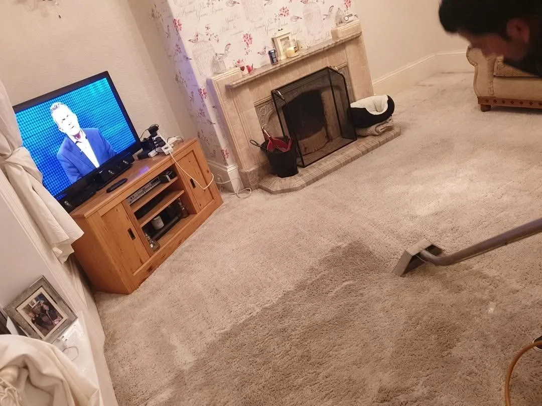 A living room with a carpeted floor, a fireplace with a protective screen, a wooden TV stand with a television showing a man in a suit, and a person vacuuming the carpet. There are some personal items and photos on furniture and shelves.
