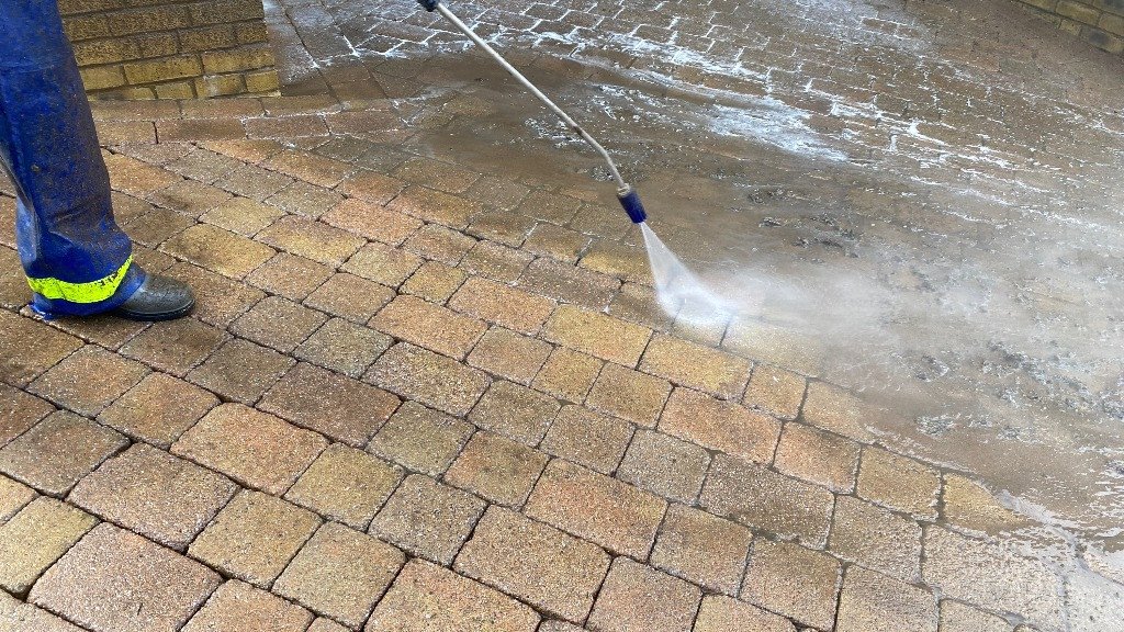 Person pressure washing a brick patio, wearing blue jeans and black shoes with yellow reflective stripes.