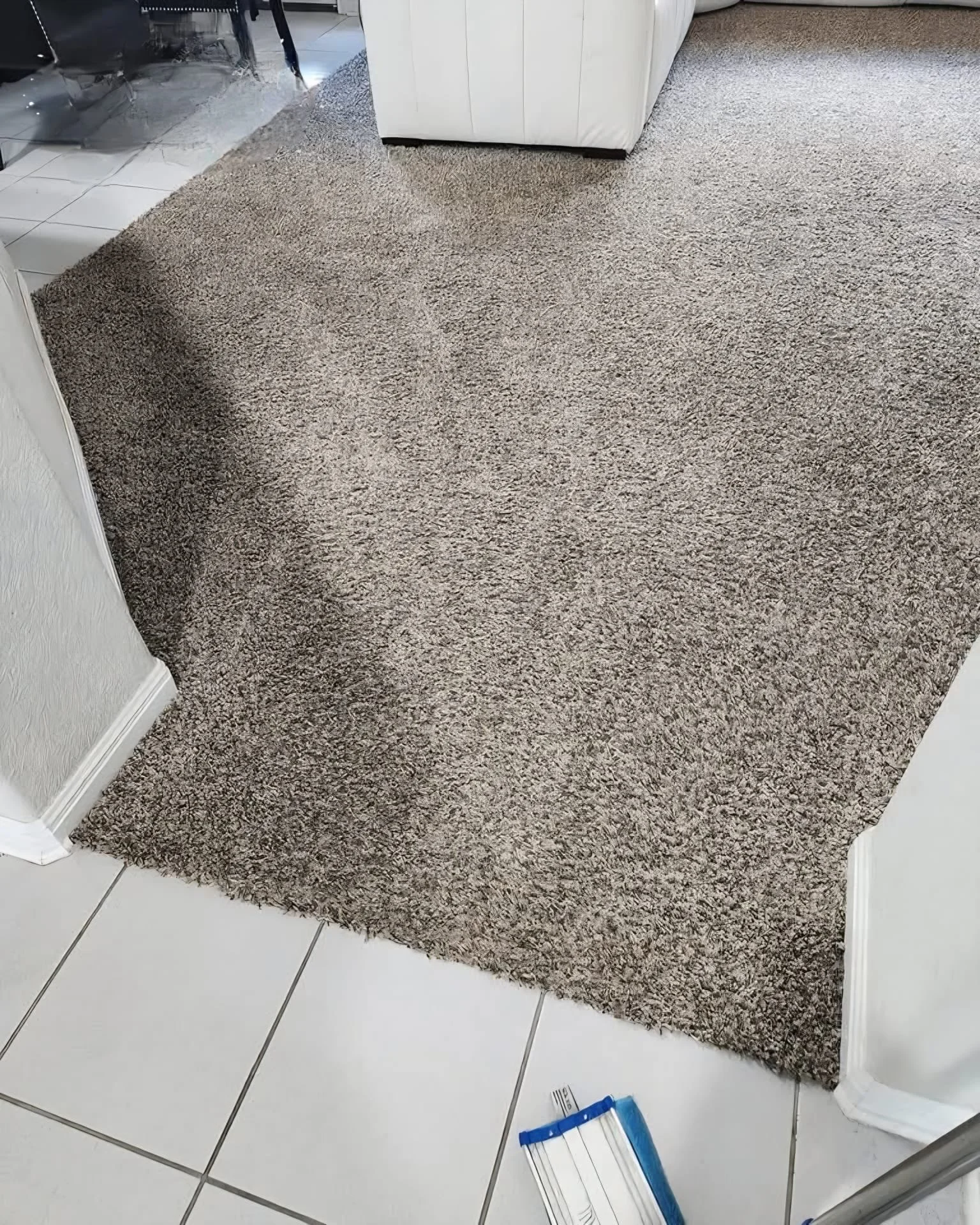 A living room corner with a beige textured area rug on white tiled flooring, white baseboards, and part of a white sofa. There is a small blue and white paint swatch or sample lying on the floor near the rug.