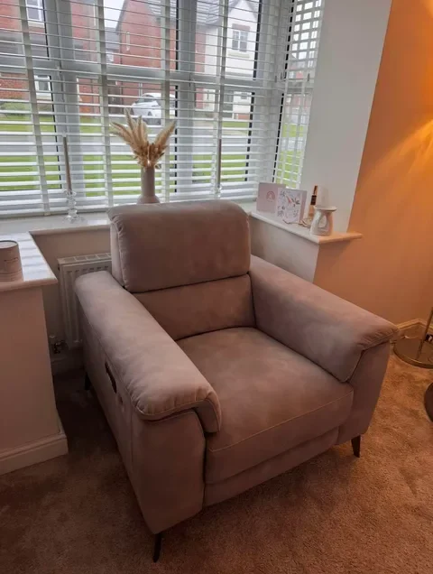 A cozy beige armchair in front of a window with white blinds, decorated with a vase of dried wheat stalks on the windowsill.