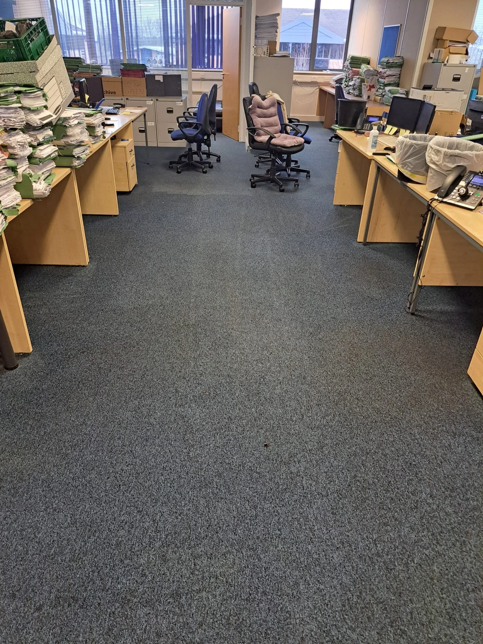 An office workspace with multiple desks cluttered with stacks of papers, filing cabinets, office chairs, and a carpeted floor, illuminated by natural light from large windows.