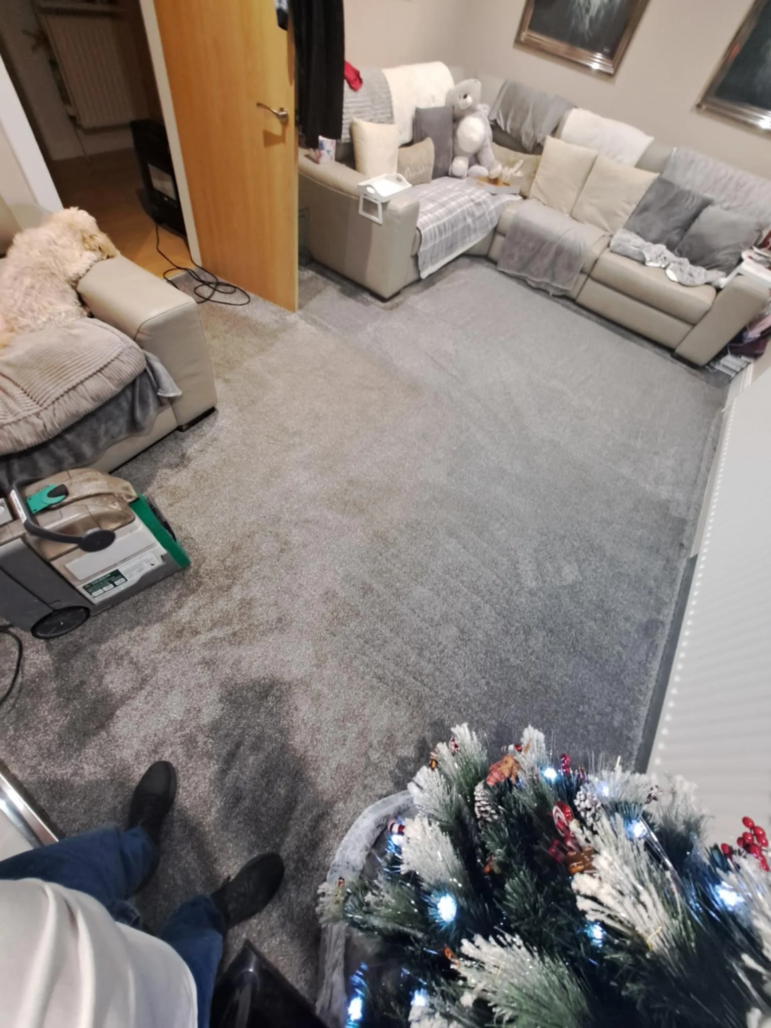 Living room with gray carpet, gray and beige couches with pillows and blankets, a Christmas tree with lights and ornaments, and a person wearing black shoes and khaki pants standing near the camera.