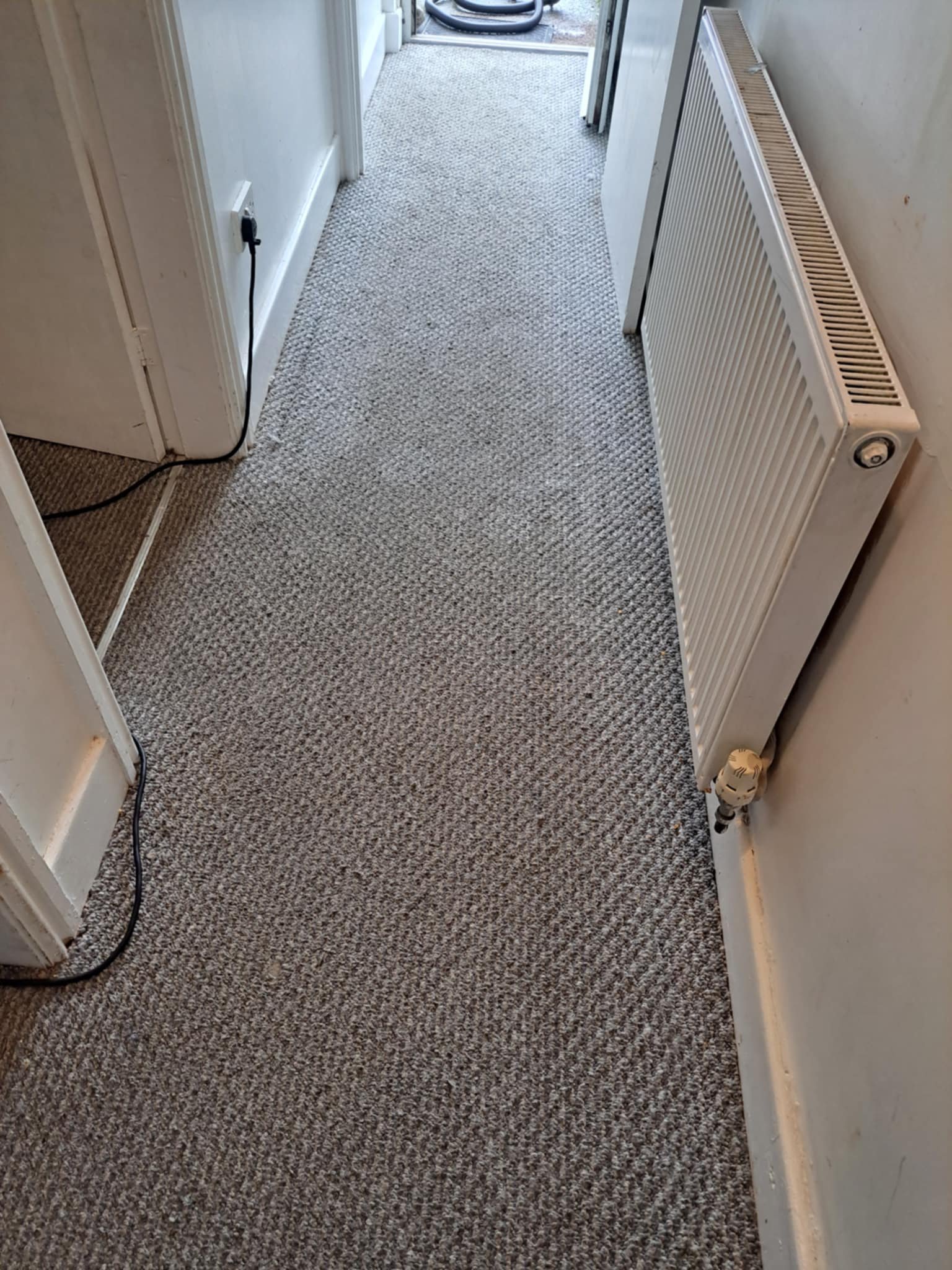 Indoor hallway with beige patterned carpet, white walls, a radiator on the right, and an open door at the end leading outside where a vacuum cleaner can be seen.