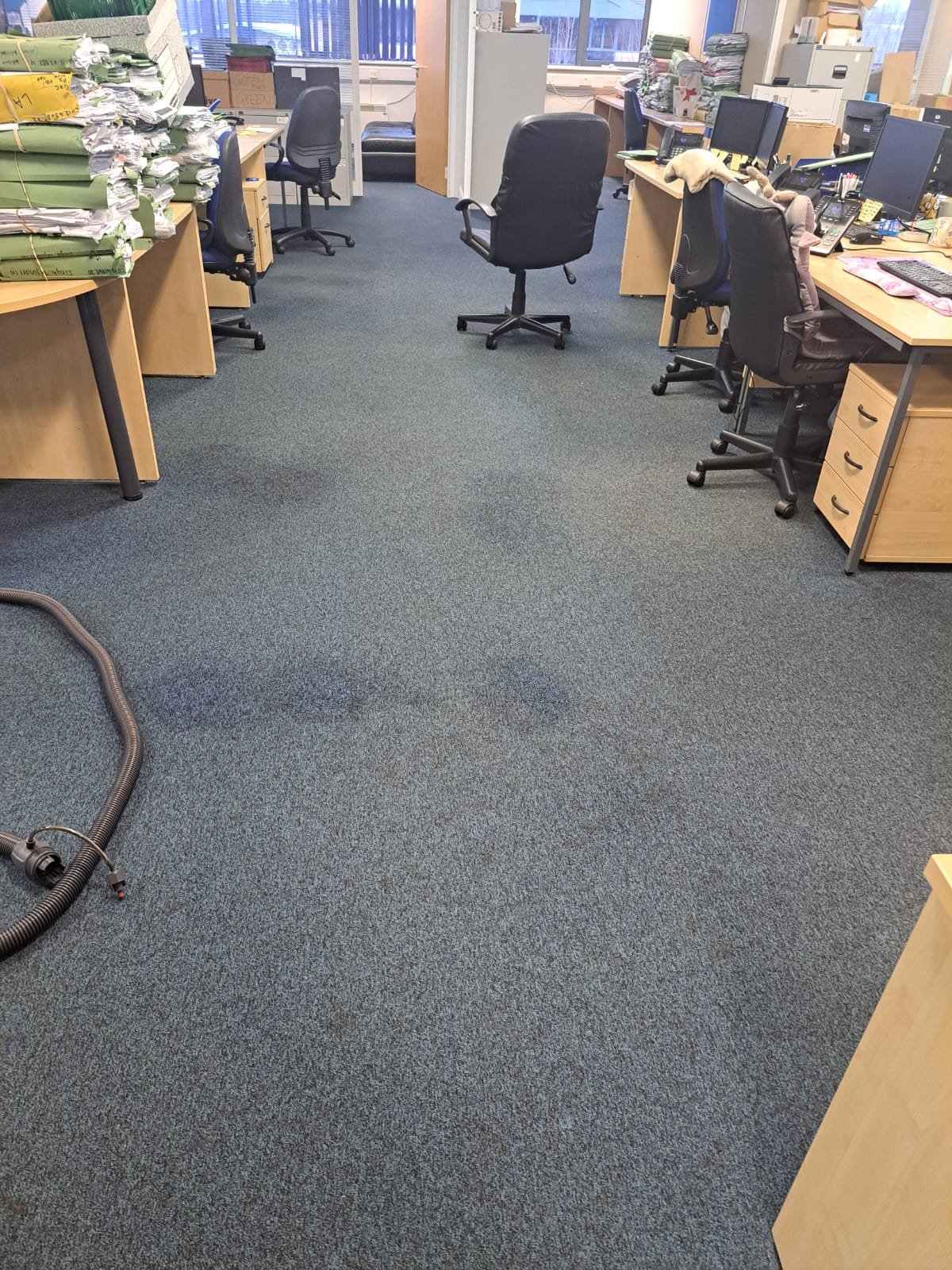 Office with desks, chairs, and computer monitors. Piles of papers and folders on desks and on the floor. A vacuum cleaner hose on the carpet. Windows with blinds in the background.