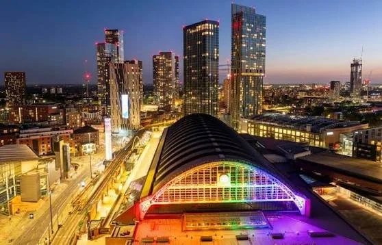 A city skyline at dusk with tall illuminated skyscrapers and a train station with a curved roof lit in rainbow colors.