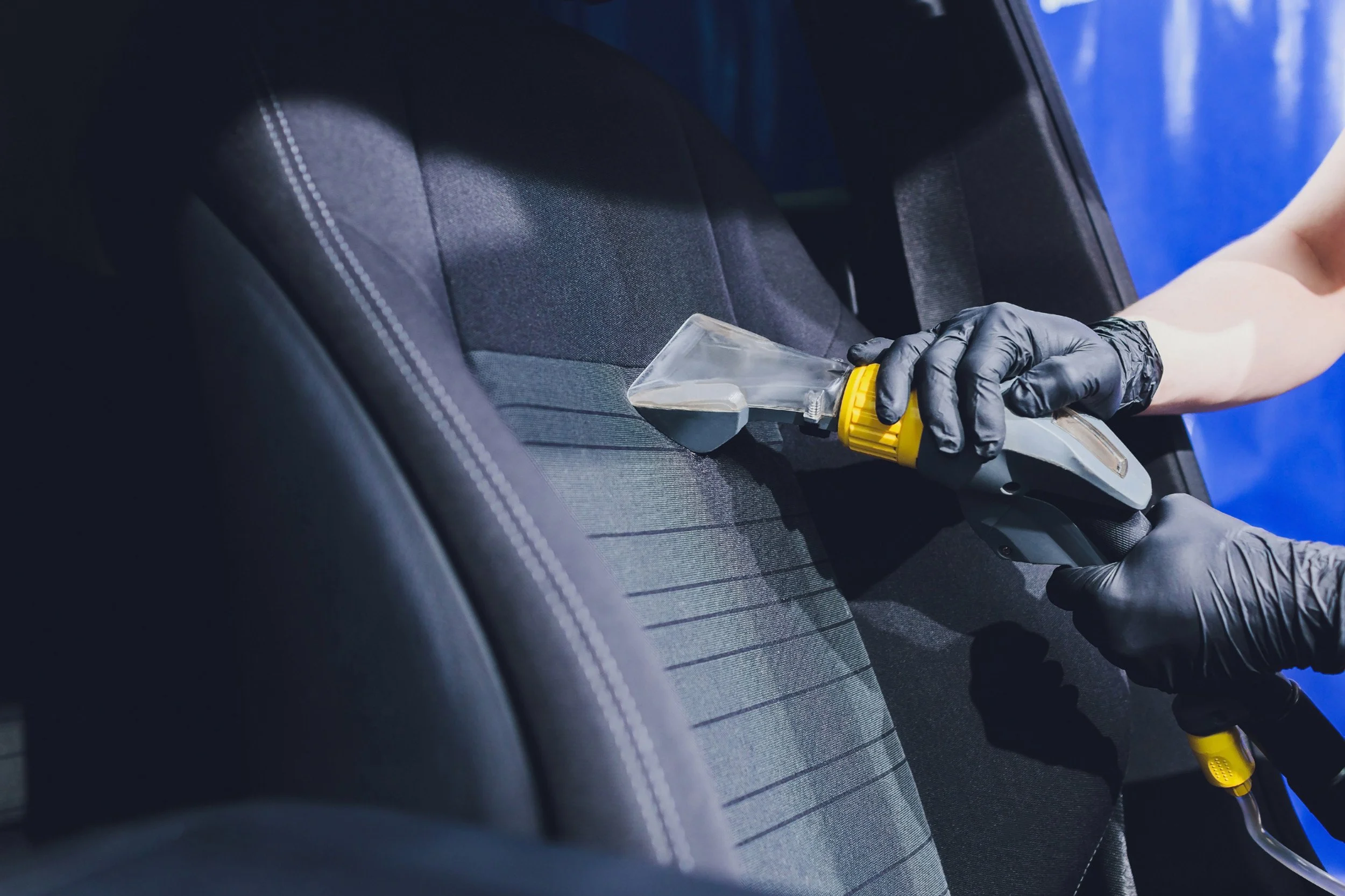 Person wearing black gloves cleaning car seat with a spray gun.