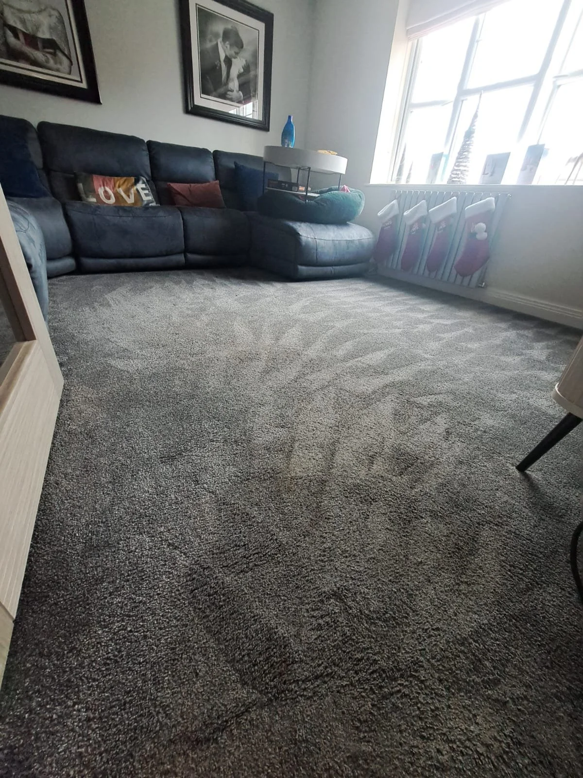 Living room with a dark leather sectional sofa, framed photos on the wall, a side table with a lamp and decorative items, a window with holiday stockings hanging, and a carpeted floor.