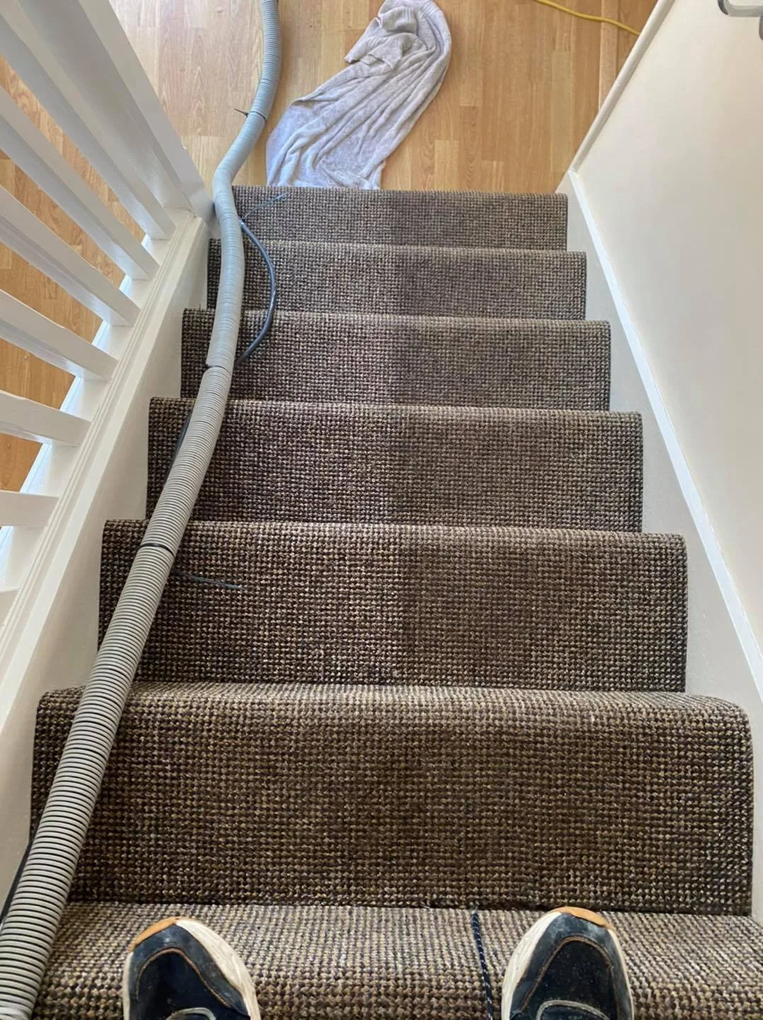 View of carpeted staircase from top, with a vacuum cleaner hose running down it, a towel at the top, and part of a person's shoes visible in the foreground.