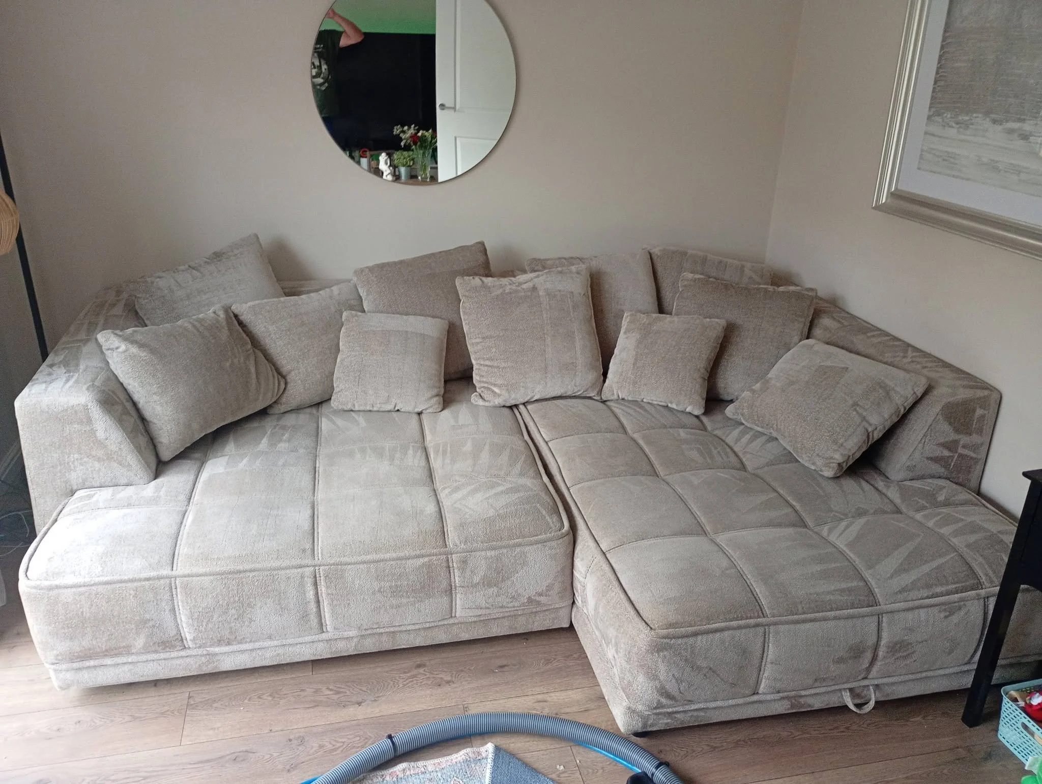 A beige sectional sofa with multiple matching throw pillows in a living room, with a mirror on the wall and a framed picture partially visible on the right side.