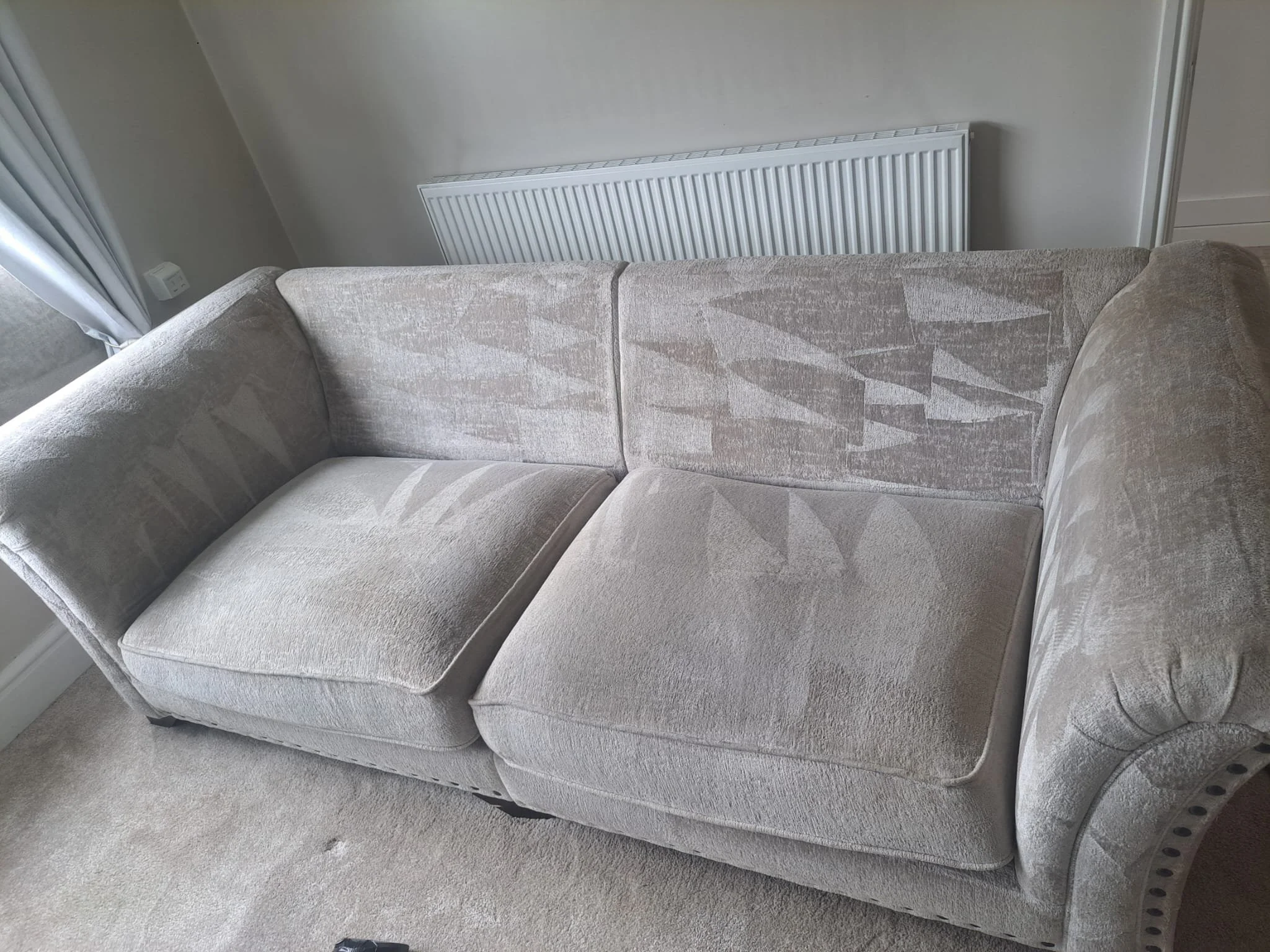 A beige fabric sofa with patterned upholstery, positioned in front of a white radiator on a light-colored carpeted floor.