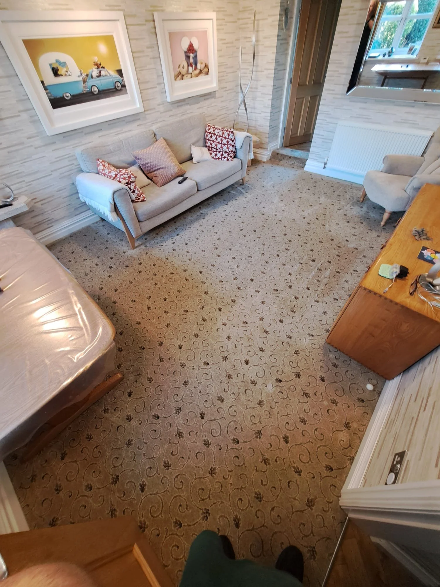 Living room with patterned carpet, beige sofa with pillows, wall art, window with garden view, wooden furniture, and person taking the photo from a high angle.