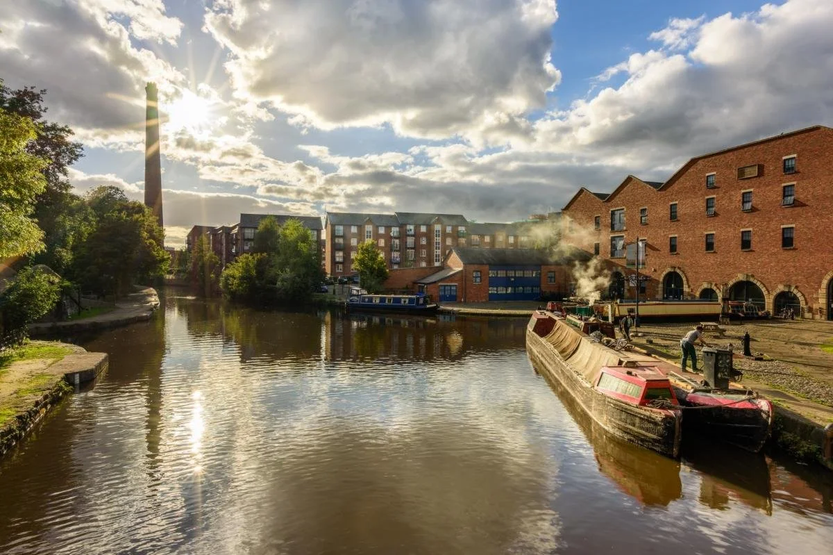 Sun shining over a canal with old brick buildings and a boat in a historic industrial area.