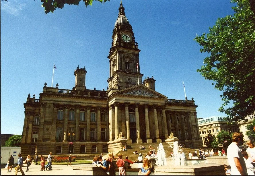 Historic city hall building with clock tower, steps, fountains, and people walking and sitting in front, under a clear blue sky.
