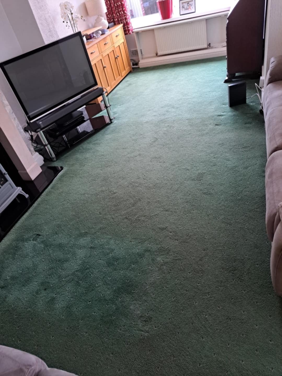 Living room with green carpet, TV on a stand, wooden cabinet, beige couch, and large window with red curtains.