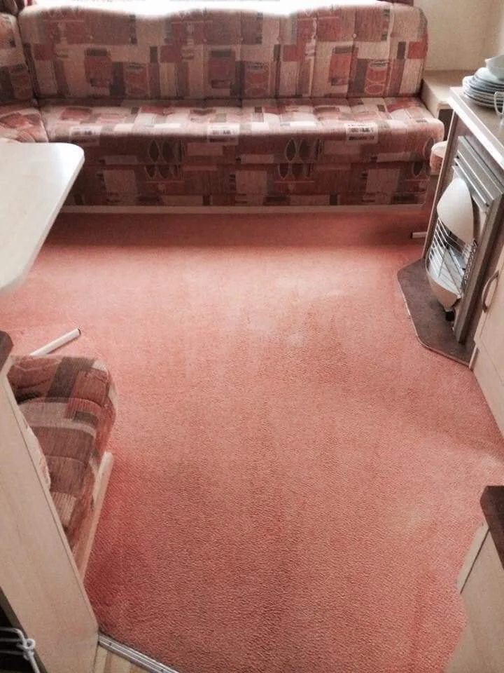 A small room with pinkish carpet, a patterned sofa along the back wall, a small sofa in the bottom left corner, a white table on the left, and a stove on the right.
