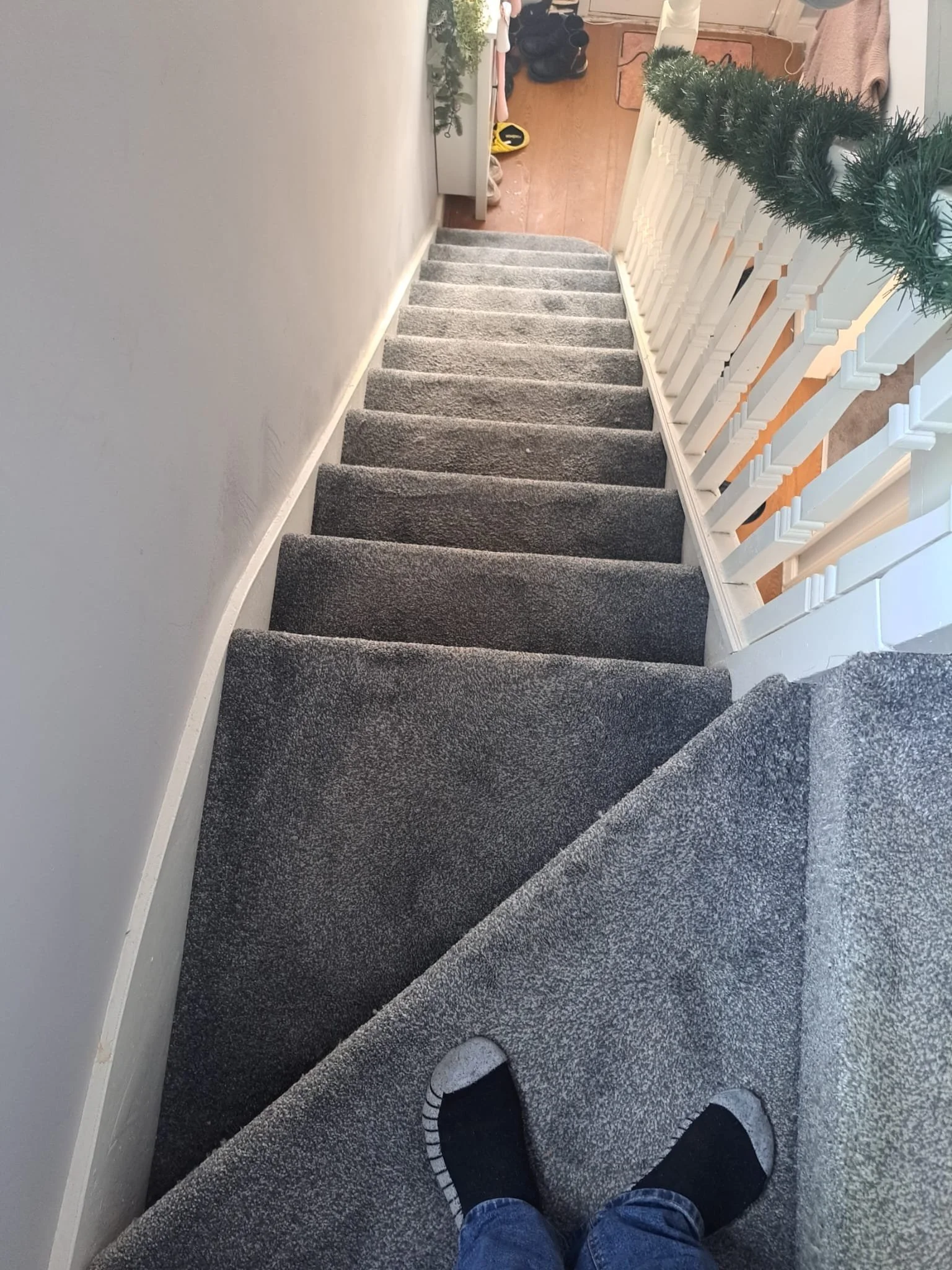 View looking down a staircase from the top, with gray carpeted steps, white walls, and a white banister. Person's legs and feet in black socks and jeans are visible at the bottom of the stairs.