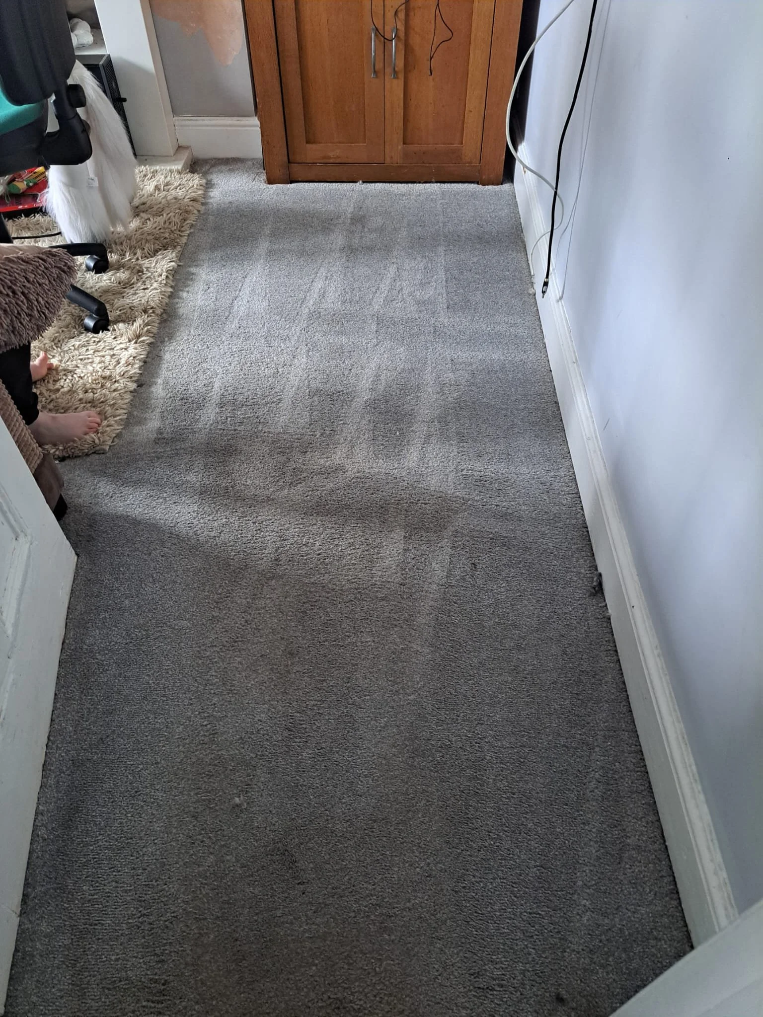 A carpeted room with a small section of a cream-colored shag rug, a wooden cabinet, some miscellaneous cords, and a chair with a furry seat cover.