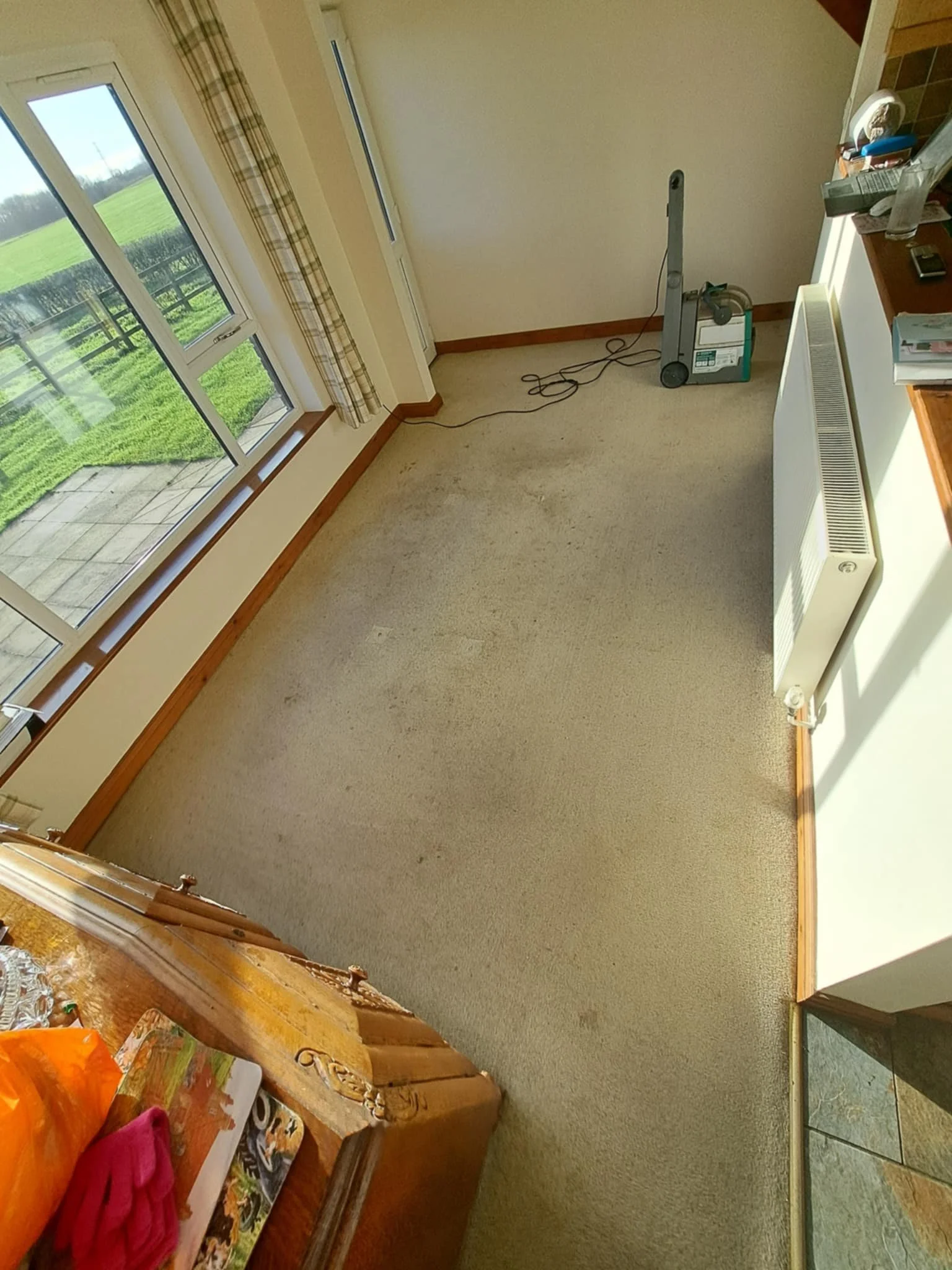 A room with large sliding glass door leading outside to an area with grass and a black fence. The room has beige carpet, a heating or cooling unit, and a wooden shelf with various items including a water bottle and magazines. There is a window with c