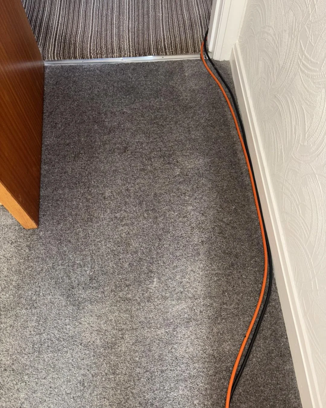 Carpeted floor in a room with two electrical cords, one orange and one black, running along the wall to a nearby door.